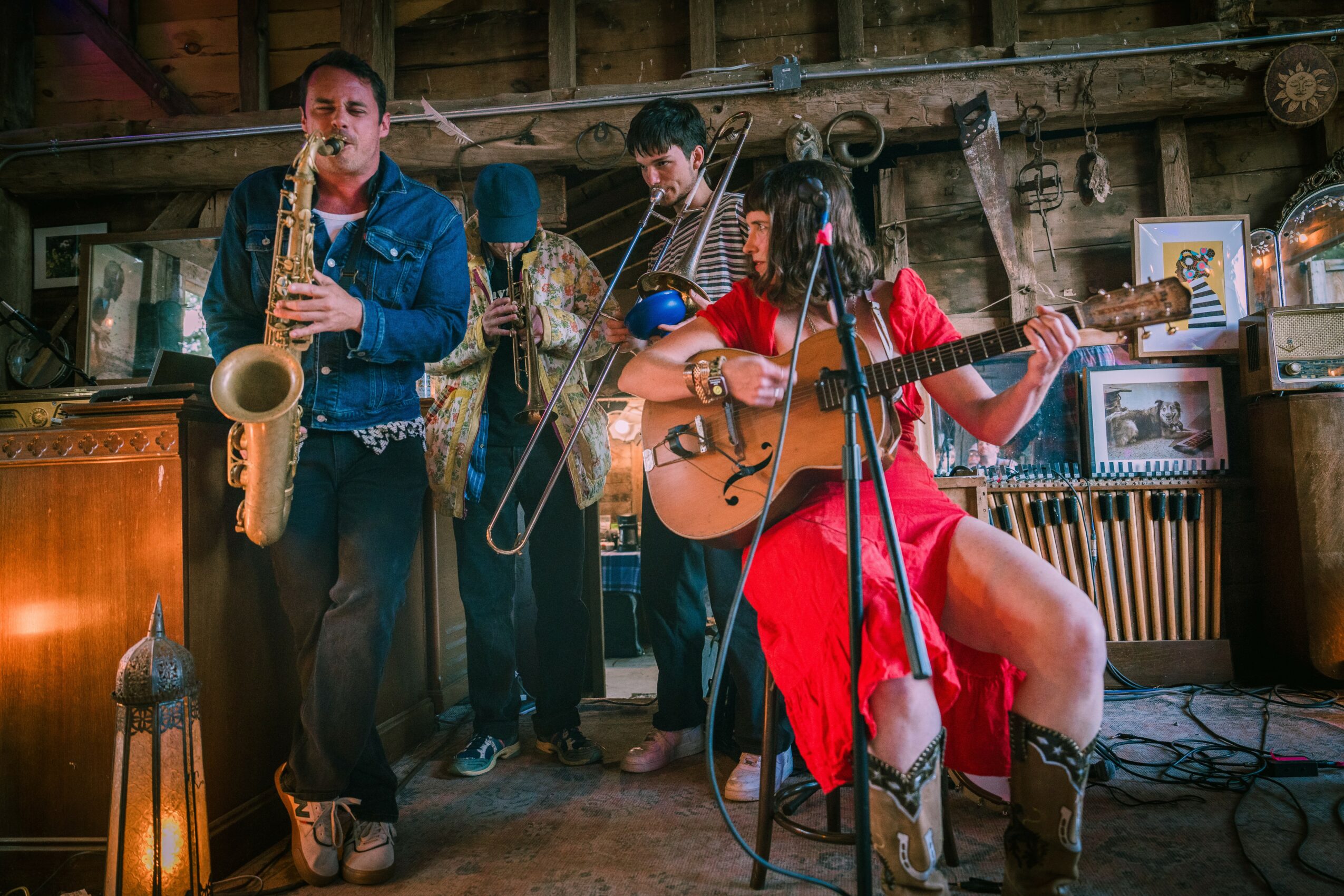 Four musicians perform in a rustic indoor setting; one plays saxophone, another on drums, a woman sings and plays guitar, all in casual attire.