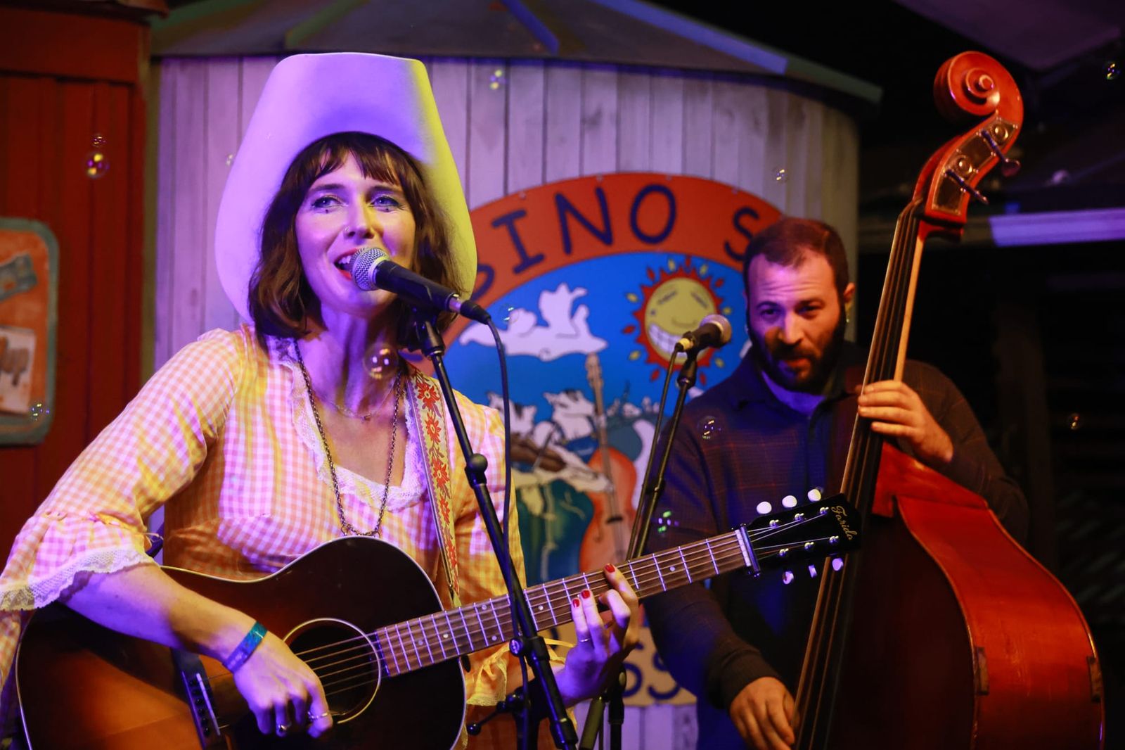A woman wearing a large white hat and yellow checkered shirt plays guitar and sings, with a man playing double bass nearby.