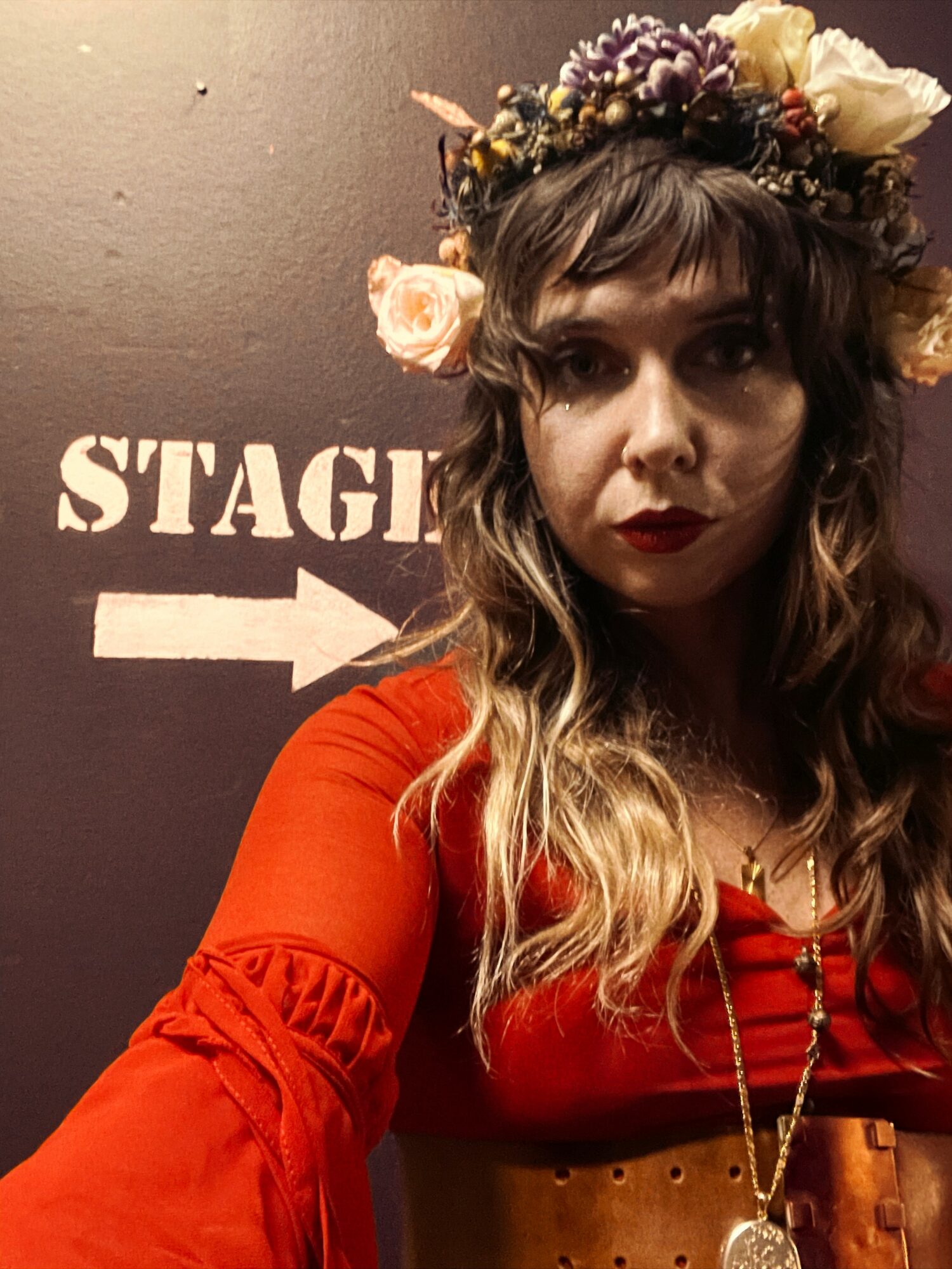 Woman with wavy hair wearing a flower crown and red dress, standing in front of a sign with an arrow.