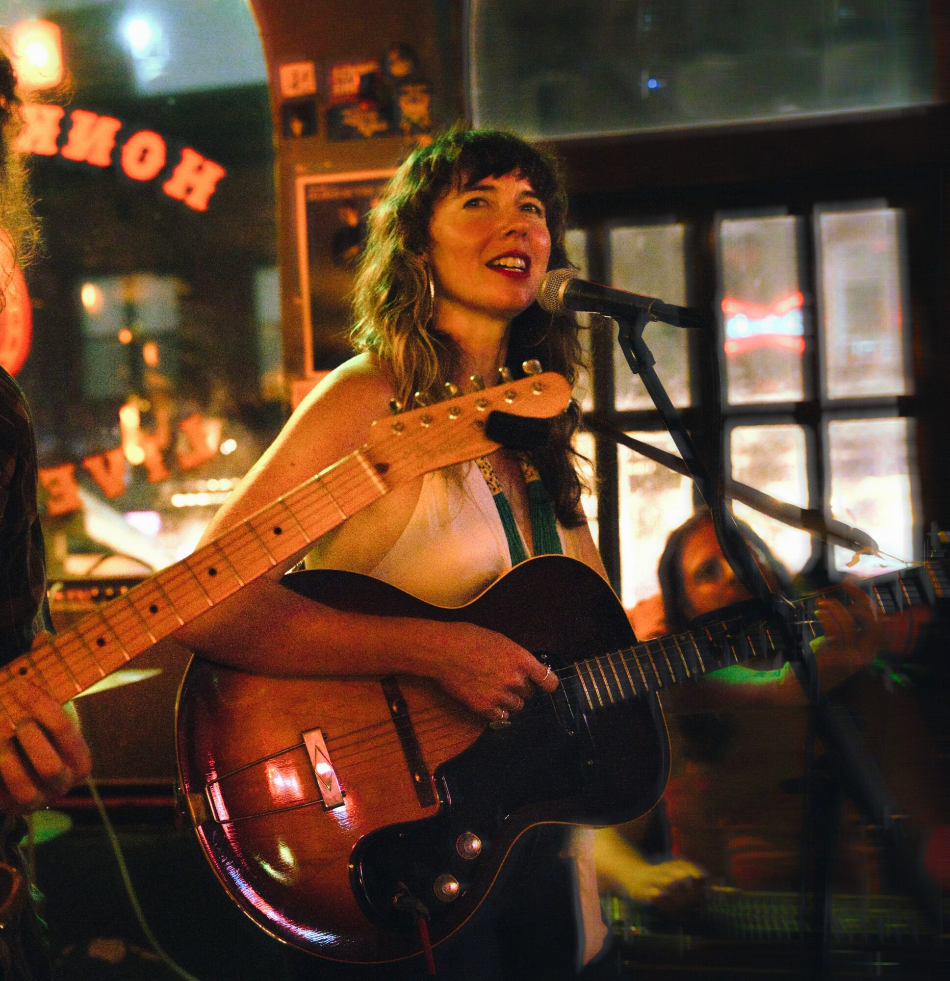 Woman singing into microphone while playing an acoustic guitar in a cozy indoor setting with warm lighting.