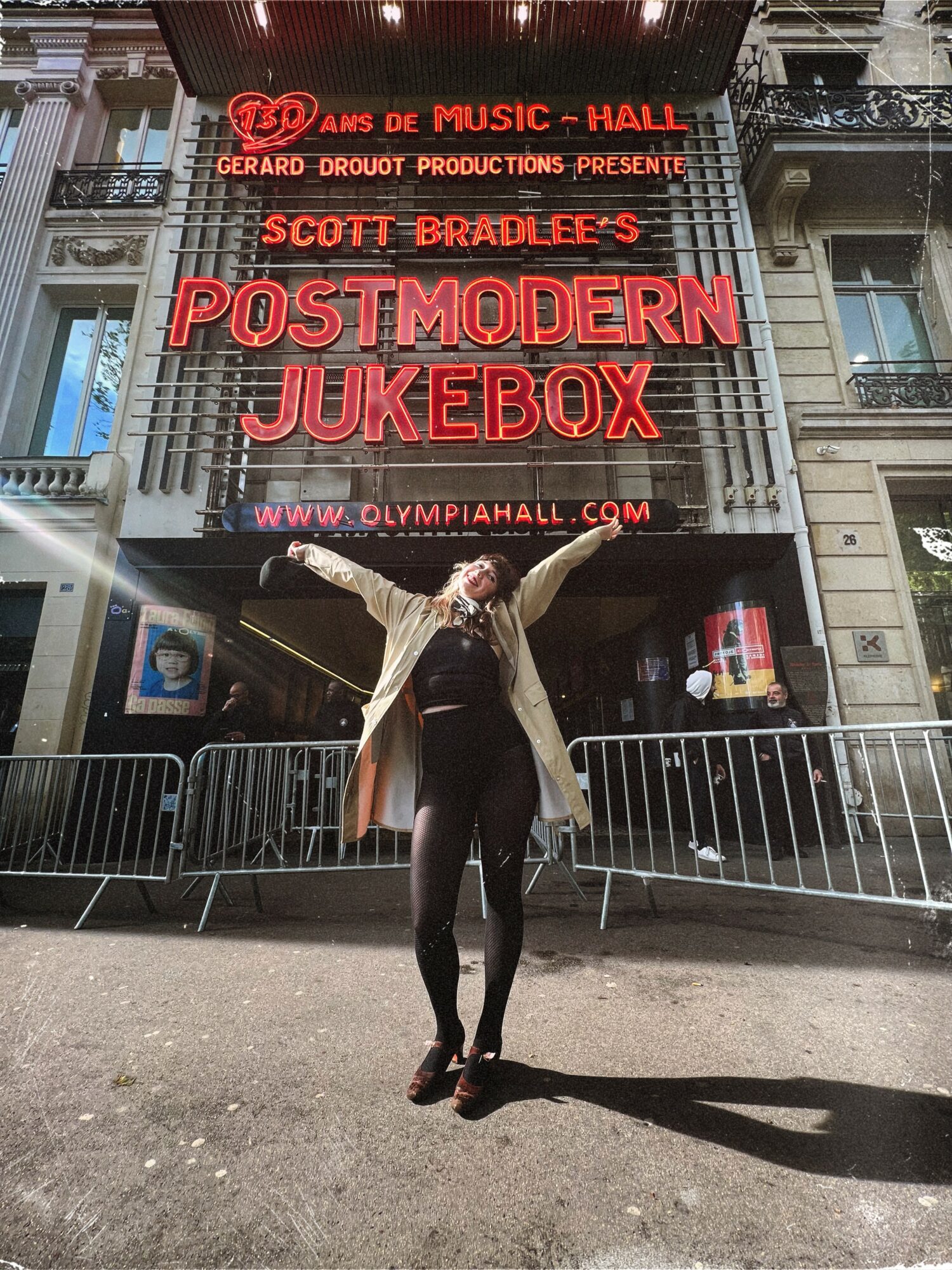 Person standing in front of a theater marquee with neon signs, arms outstretched, on a city street at night.