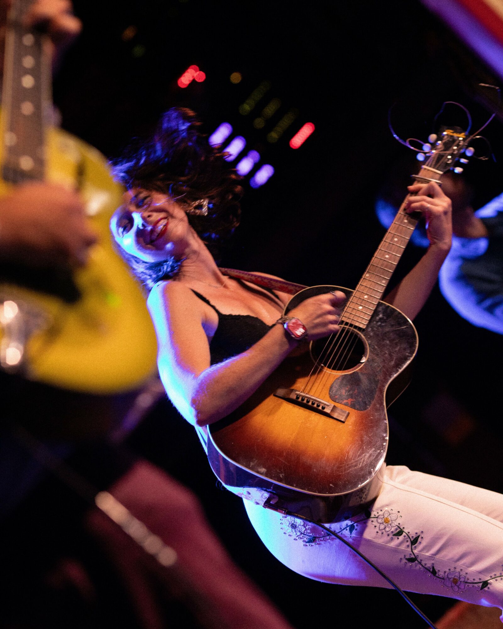 Woman playing an acoustic guitar on stage, smiling, with colorful lights in the background.