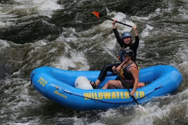 Two people in a blue inflatable boat navigating white water rapids, one person paddling and the other holding an oar above head.