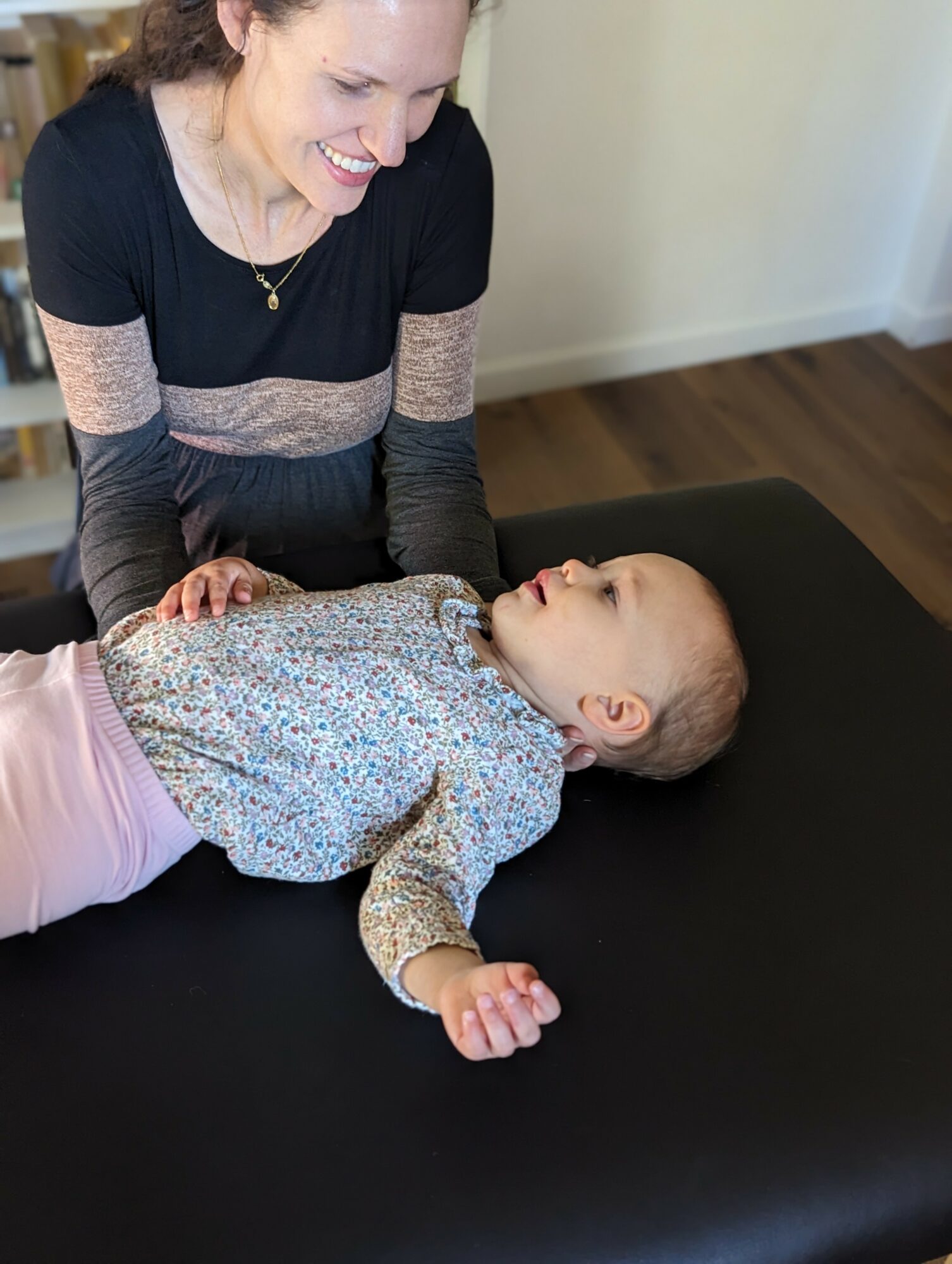 Woman smiling at a young girl lying on a black table, indoors with wooden floor and white wall in background.