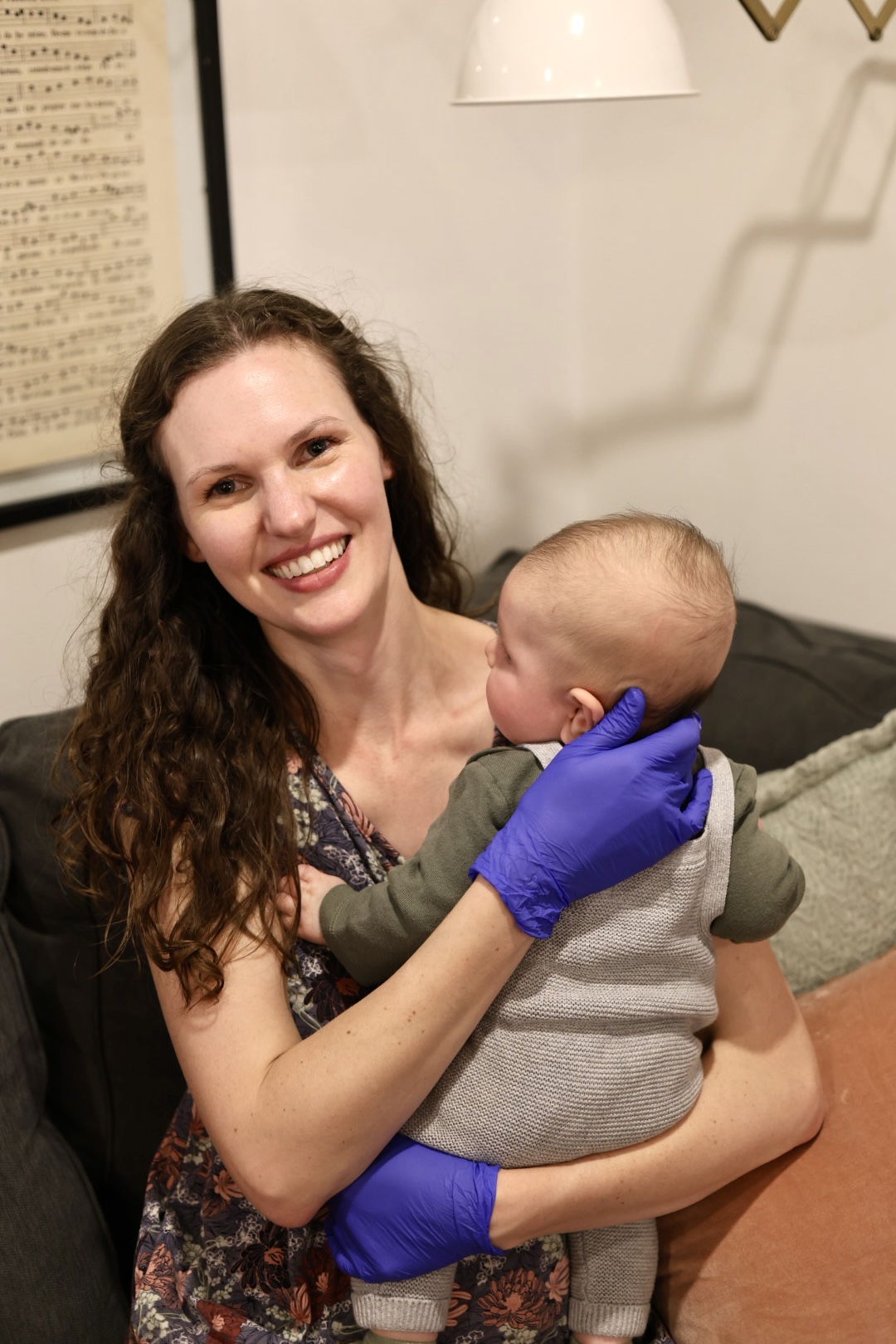 Woman with long curly hair holding a baby, both wearing blue gloves, smiling indoors.