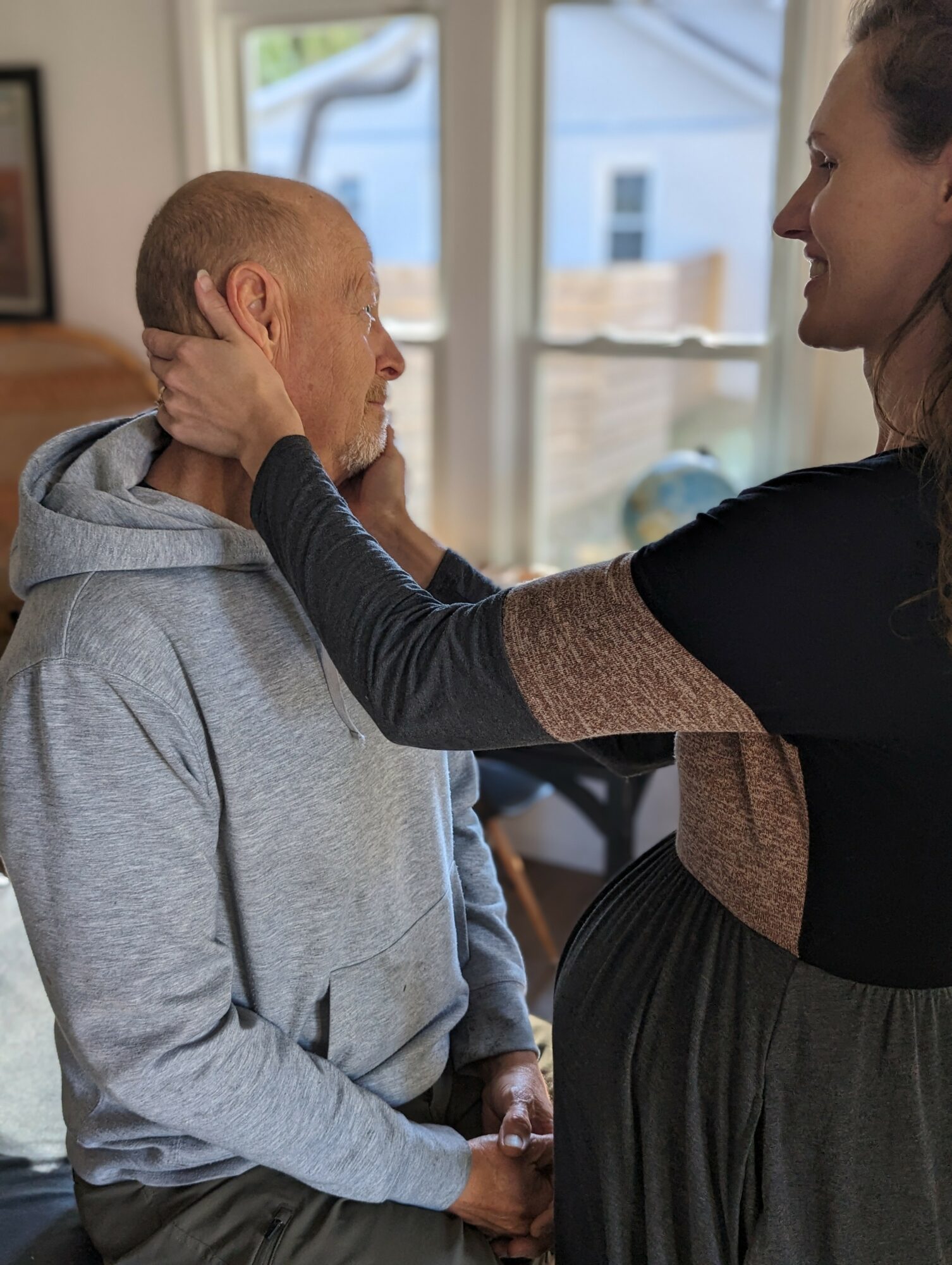 Woman gently holds man's face, both looking at each other indoors near a window.