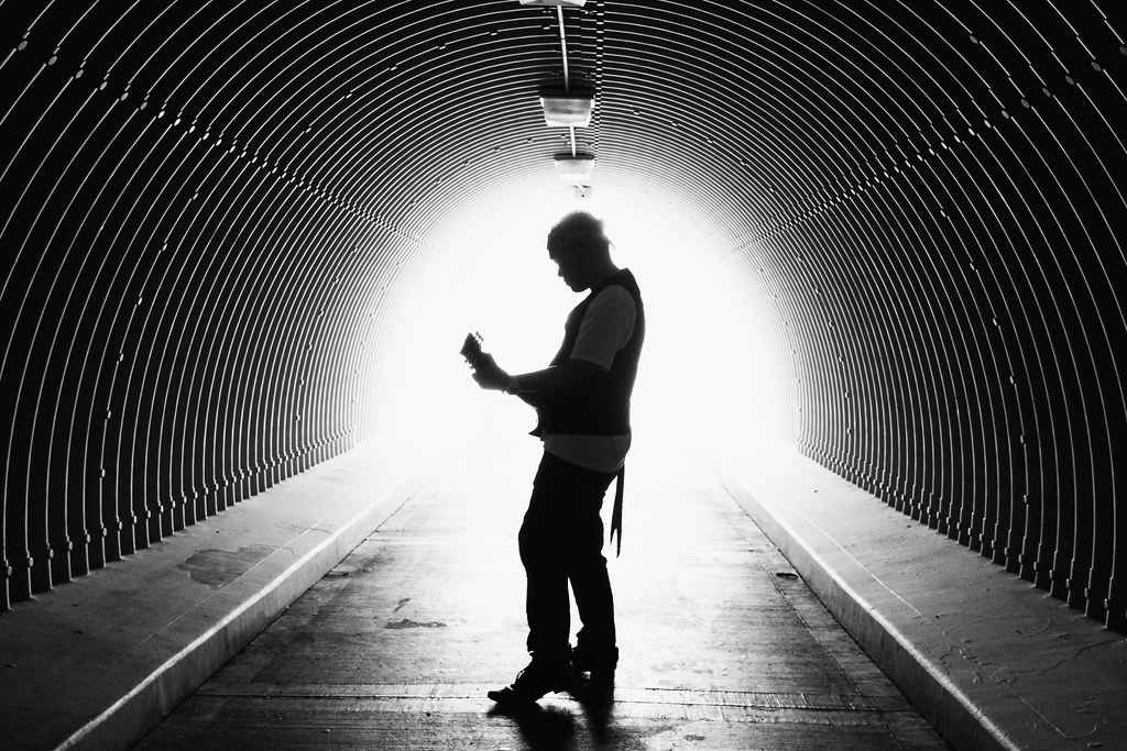 Silhouette of a person standing in a tunnel, looking at a device, with bright light at the tunnel's exit behind them.