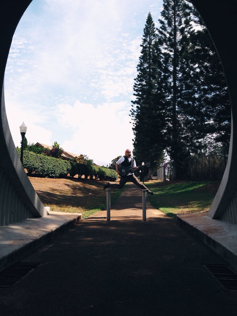Person standing on a pathway with trees and sky in the background, viewed from inside a tunnel or archway.