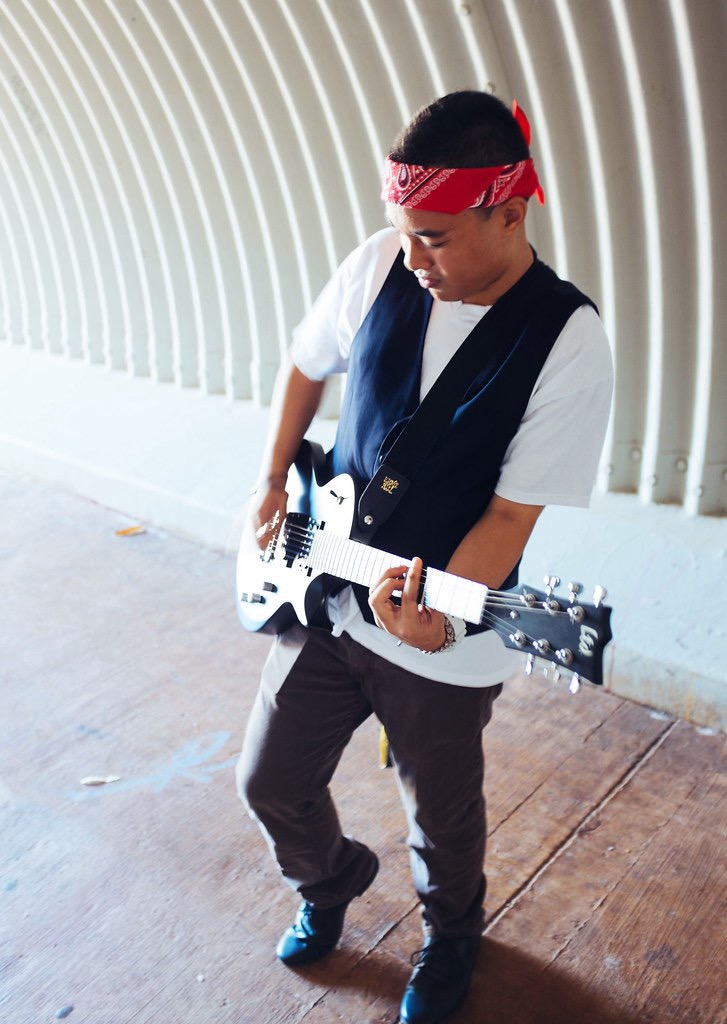 Young person wearing a red bandana, playing a white electric guitar, standing against a curved wall with vertical bars.