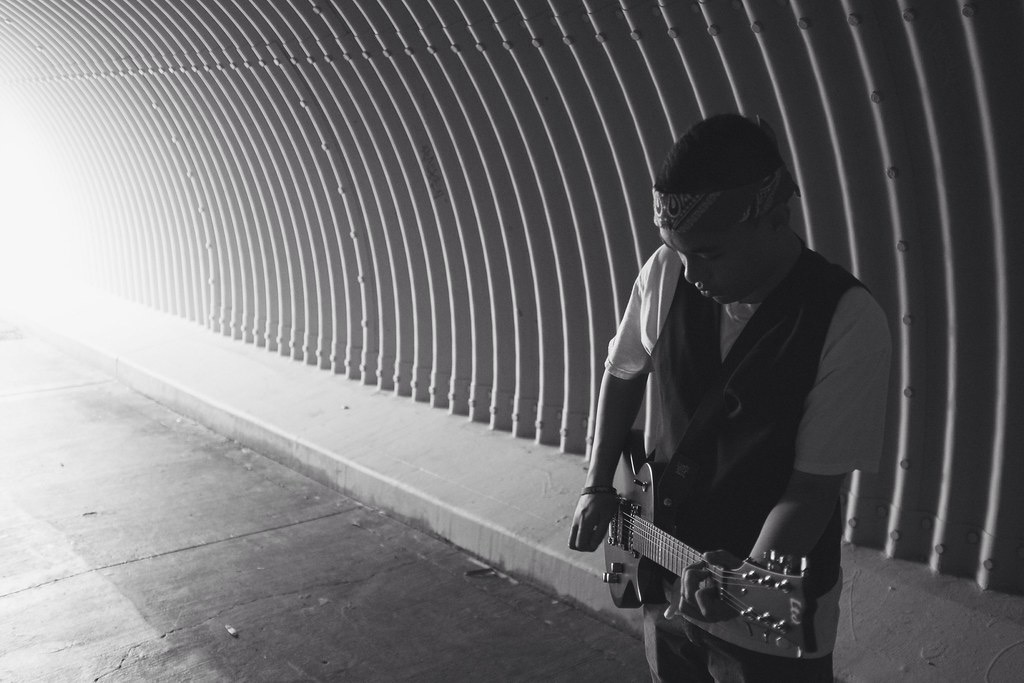 Person playing guitar in a tunnel with curved walls, black and white photo, sunlight at the tunnel's entrance, person wearing glasses and a vest.