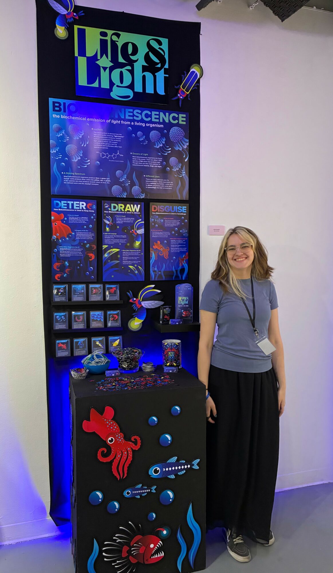 Girl standing next to a colorful science exhibit with glowing elements and a large display screen.