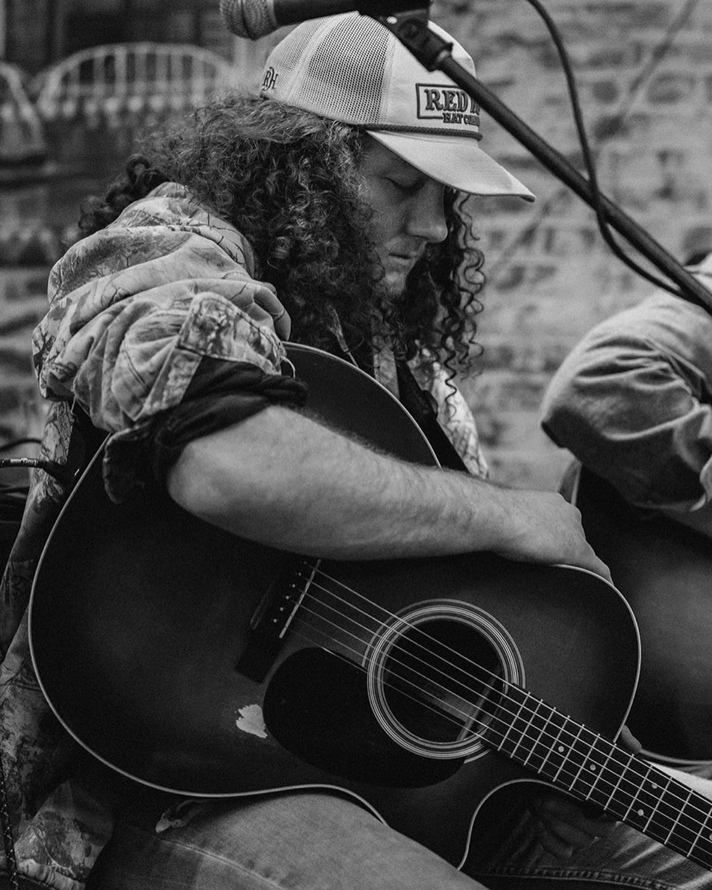 Person with curly hair wearing a cap, playing an acoustic guitar outdoors, with a microphone overhead, in black and white.