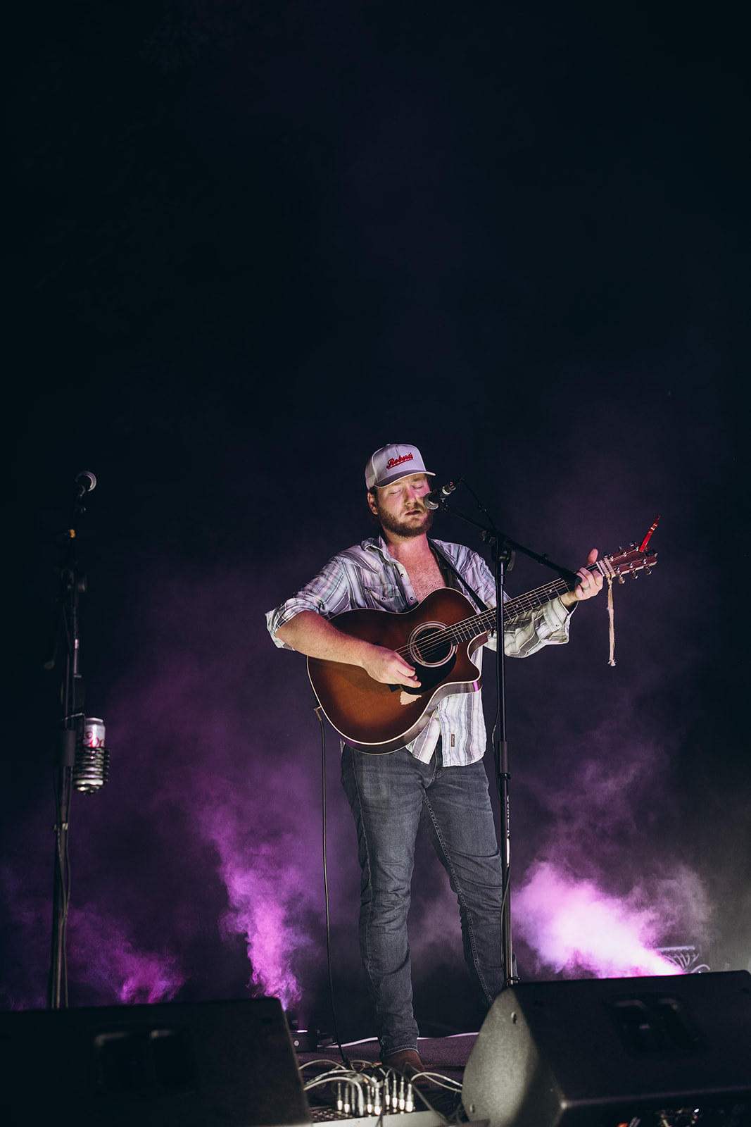 Person playing guitar and singing on stage with purple lighting and fog, wearing a hat and casual clothes.