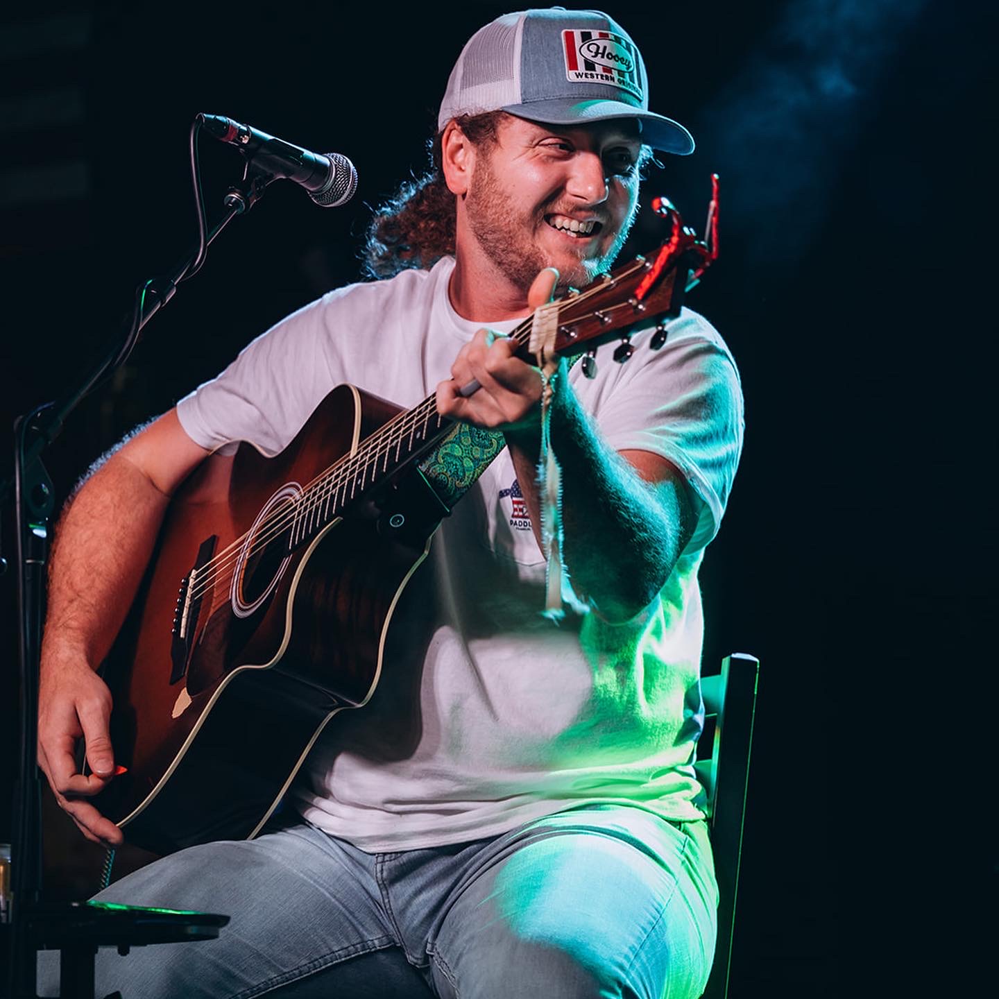 Person playing guitar and singing into a microphone on stage, smiling, wearing a cap and casual clothes.