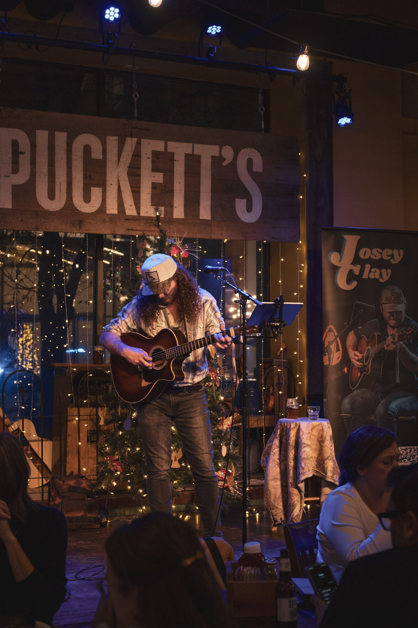 Musician playing guitar on stage with a sign reading 'PUCKETT'S' behind, decorated with string lights, in a dimly lit venue.