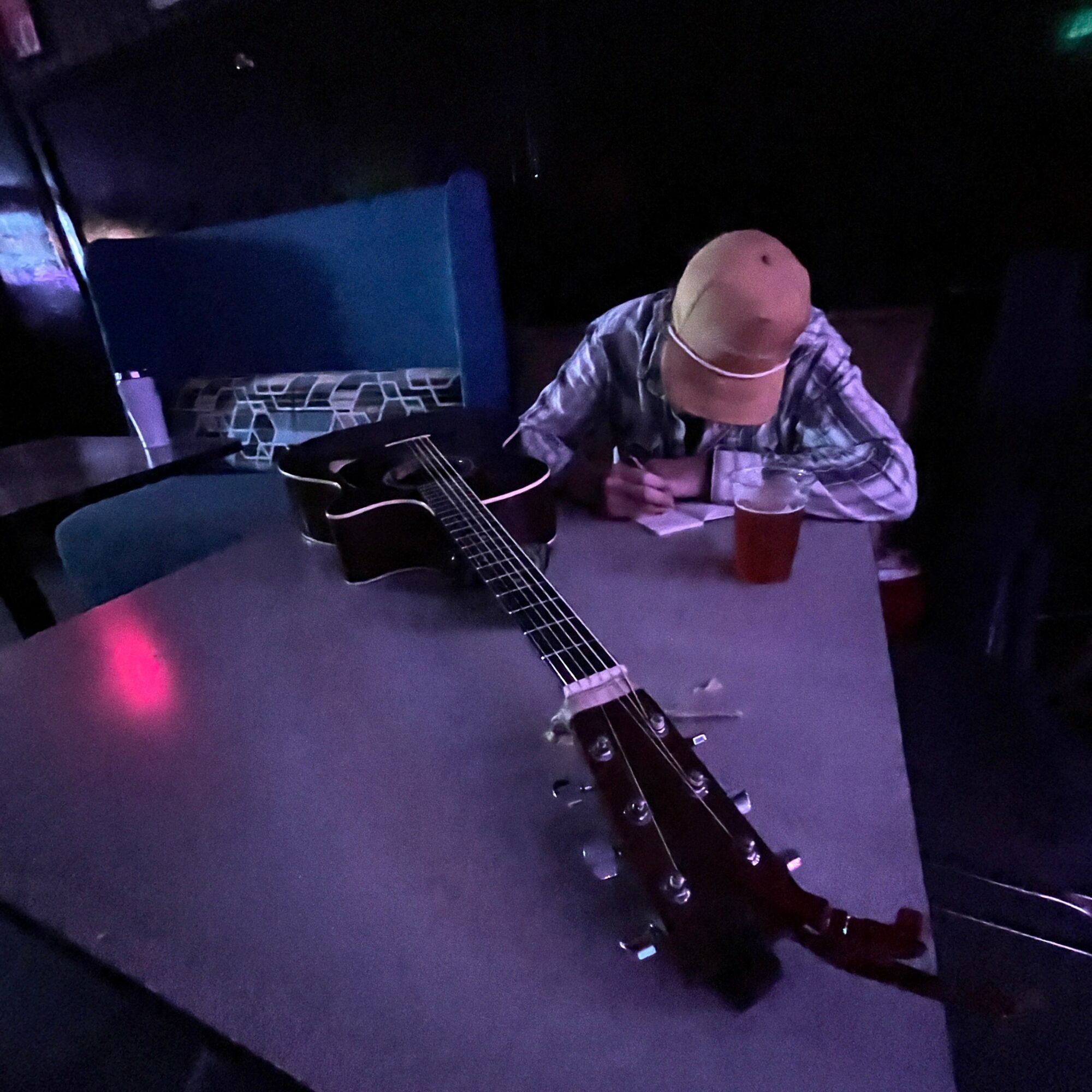 Person wearing a hat sitting at a table with an acoustic guitar and a drink, writing or drawing on paper.