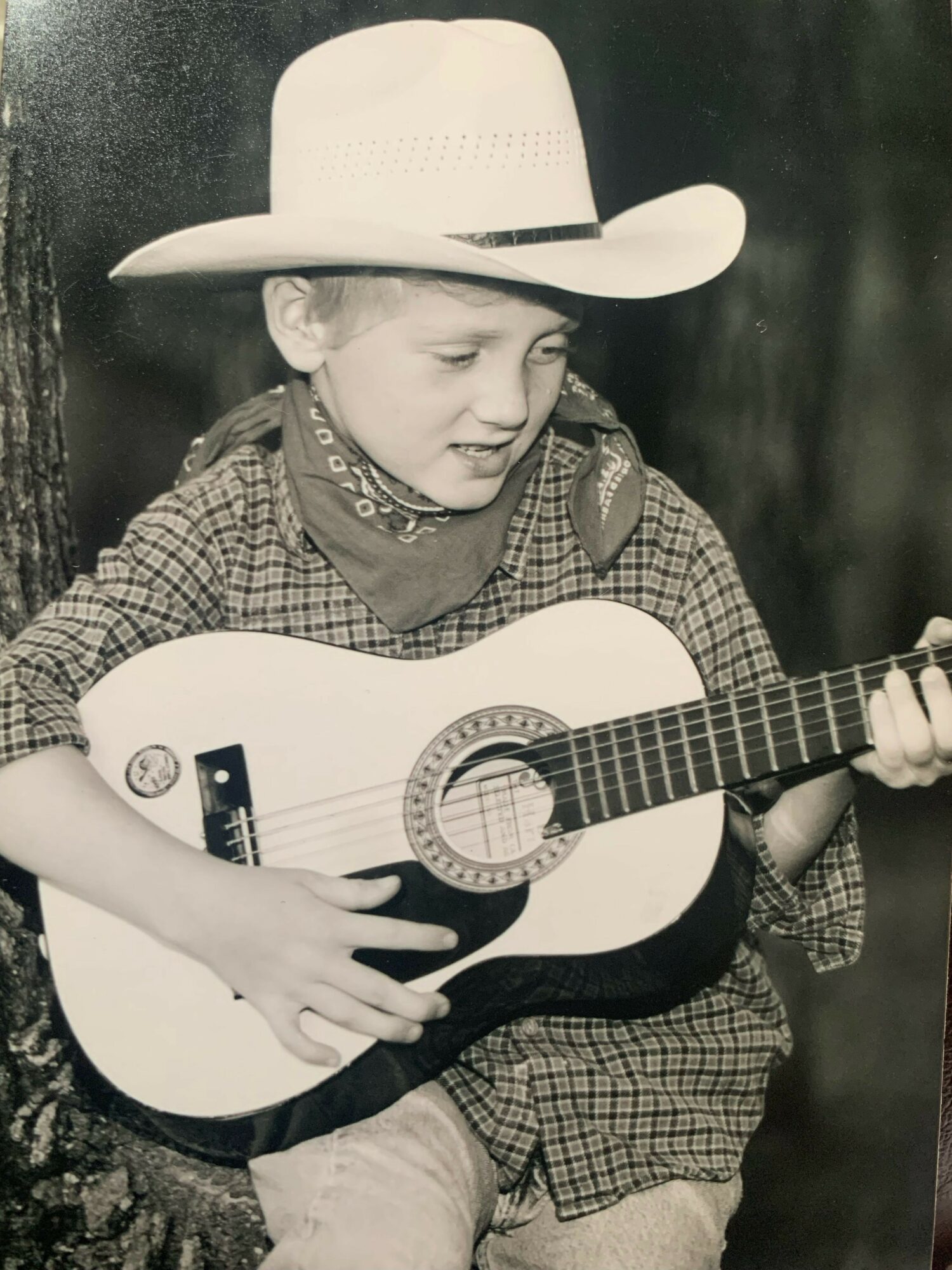 Child wearing a hat and bandana playing an acoustic guitar, lying down outdoors.