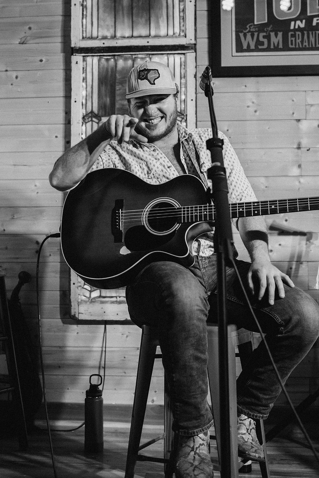 Person playing guitar and singing into microphone on stage, wooden background, smiling, wearing a cap, casual clothing.