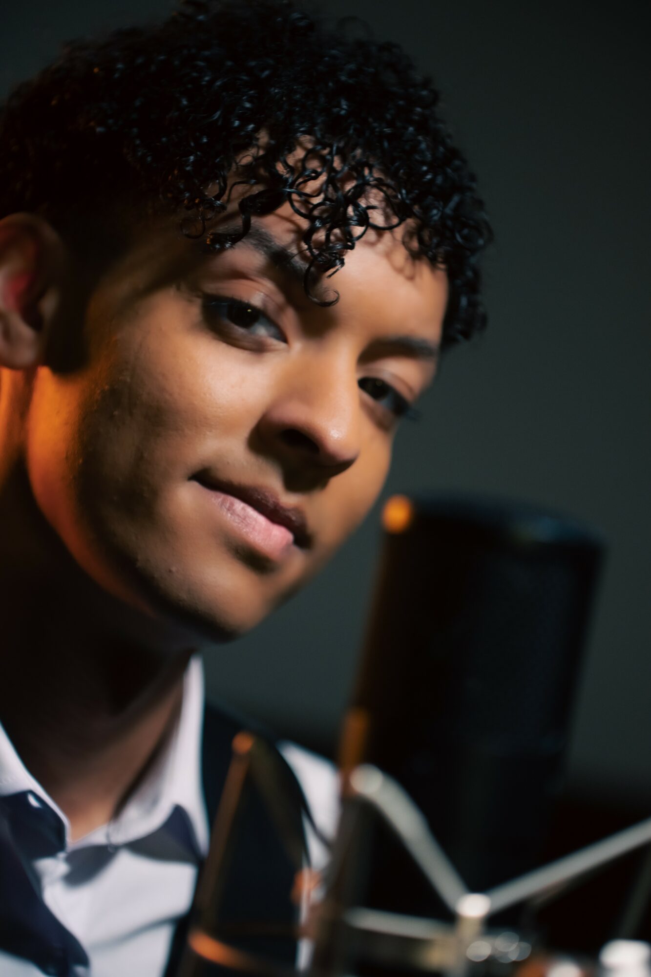 Young person with curly hair smiling, holding a smartphone, in a dimly lit environment.