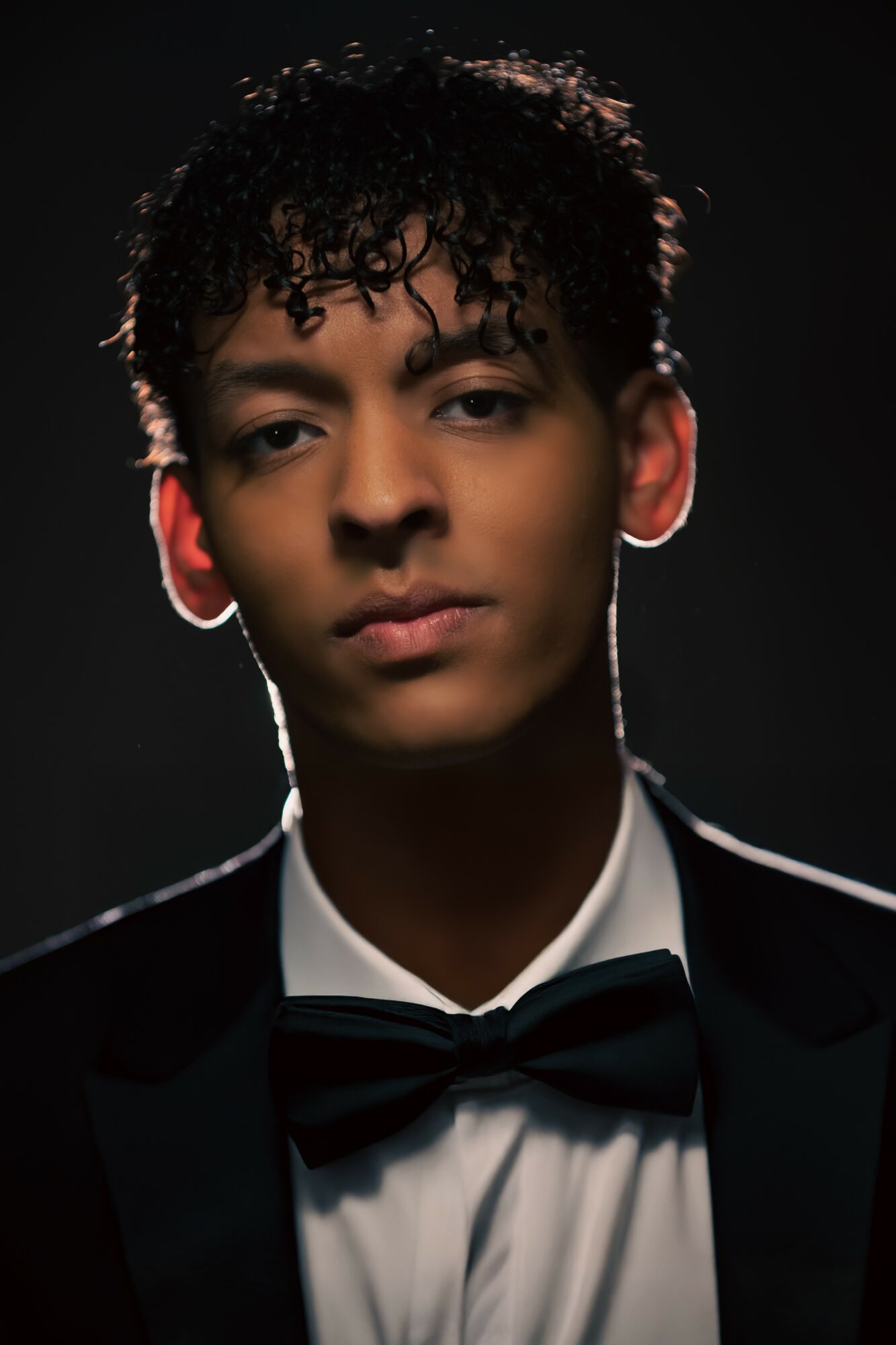 Young man with curly hair in formal attire, dark background, serious expression, illuminated from behind, tuxedo and bow tie.
