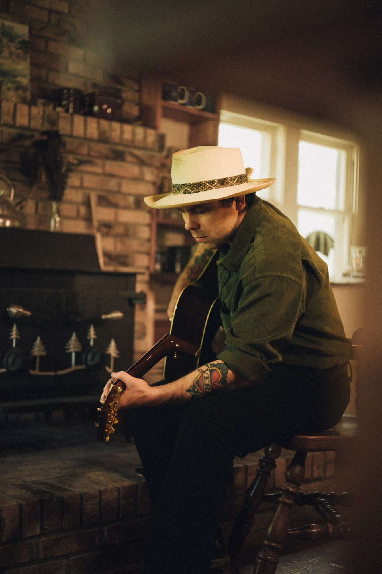 Person wearing a wide-brimmed hat and green jacket playing guitar indoors near a brick fireplace and windows.