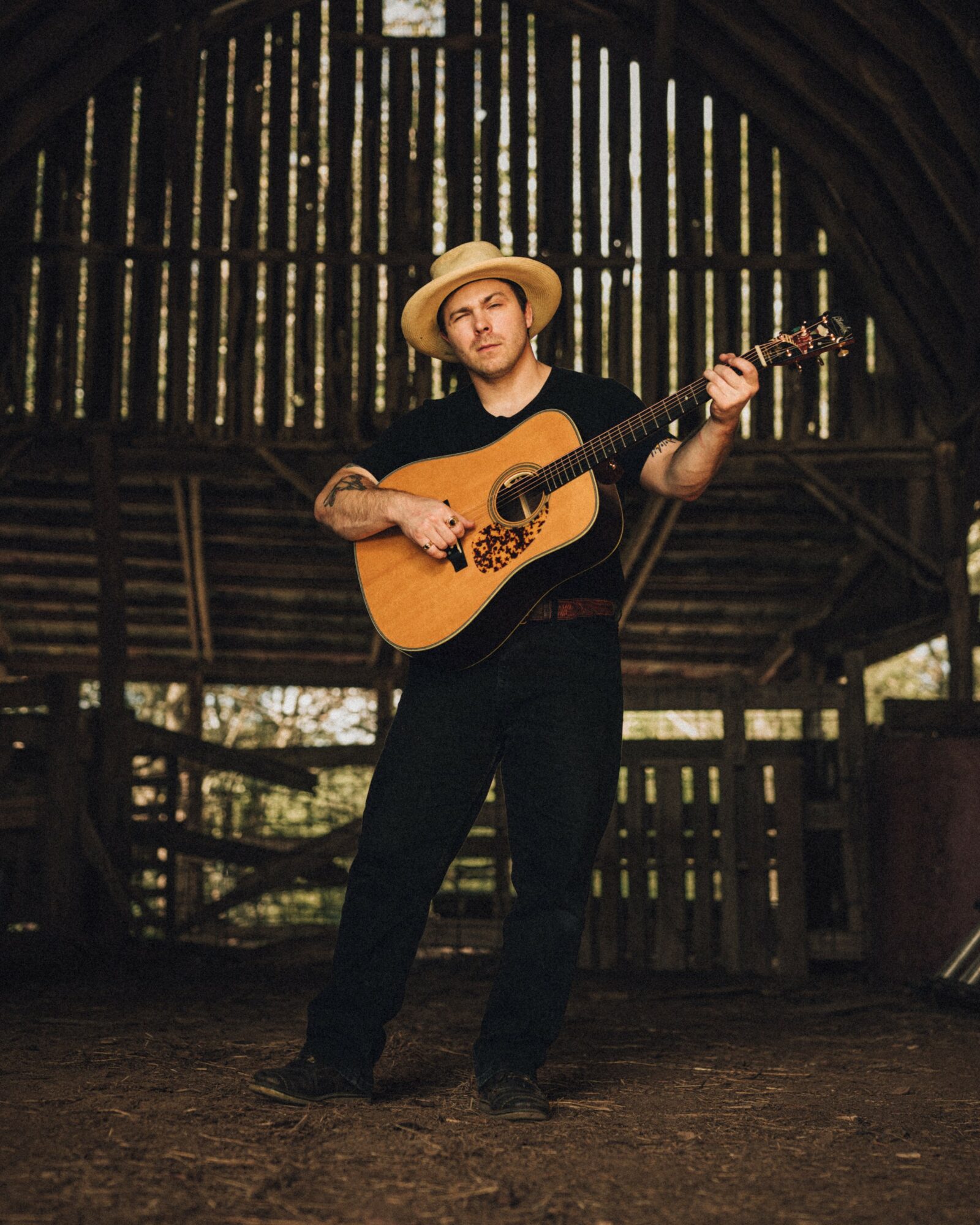 Person wearing a straw hat playing an acoustic guitar inside a wooden structure with vertical slats.