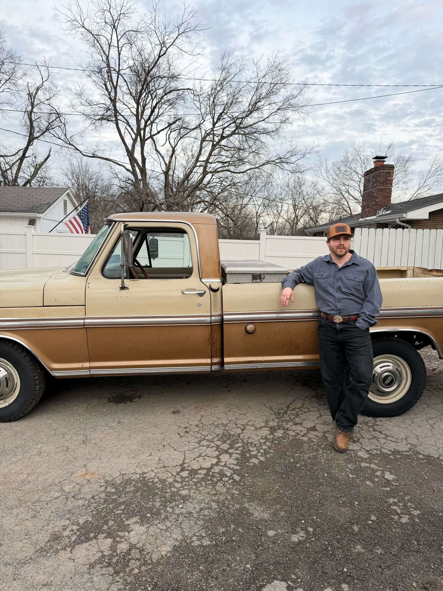 Man standing next to a vintage beige pickup truck parked on a street, with a leafless tree and buildings in background.