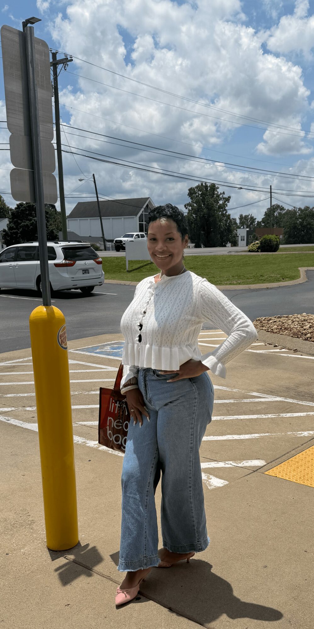 Woman standing outdoors in a parking lot, smiling, with a cloudy sky and trees in the background.