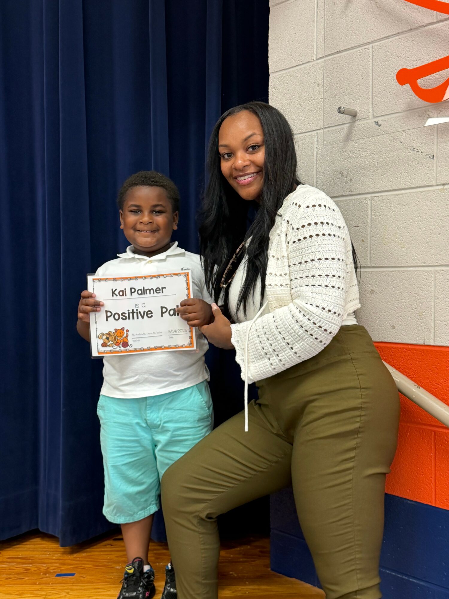 Young girl holding a certificate with a woman smiling beside her, standing in front of a blue curtain.