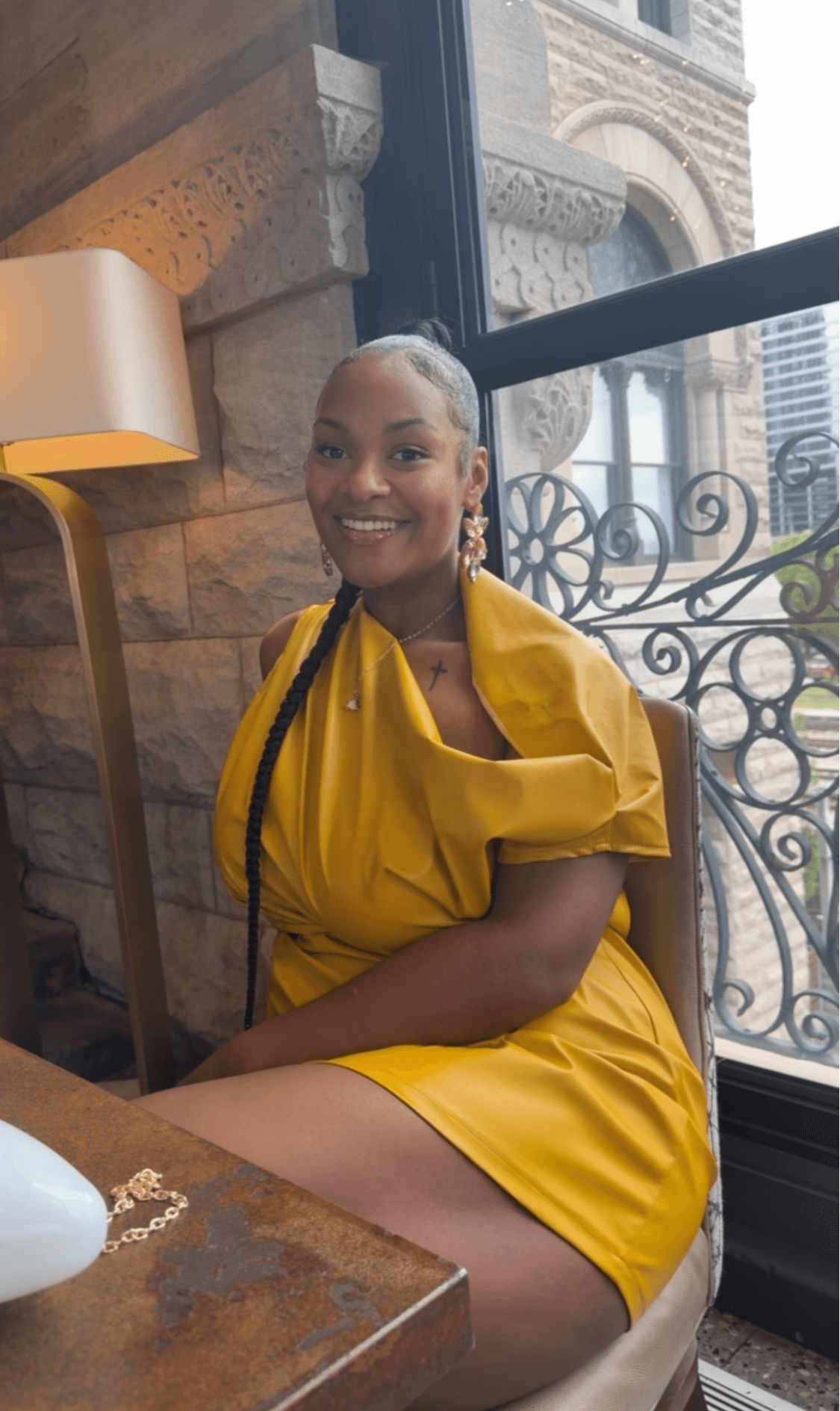 Smiling woman in yellow dress sitting by window with ornate railing and cityscape background.