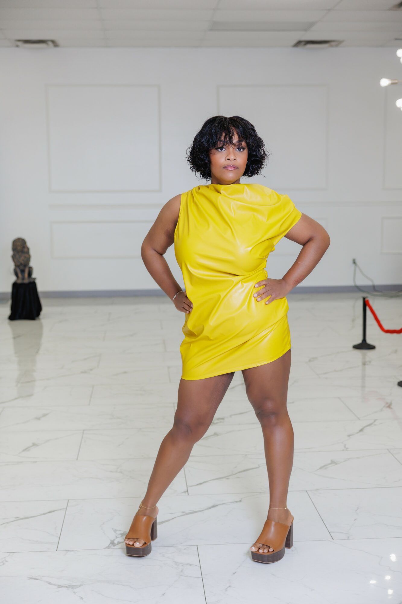 Woman in yellow dress standing with hands on hips in gallery with white walls and marble floor.