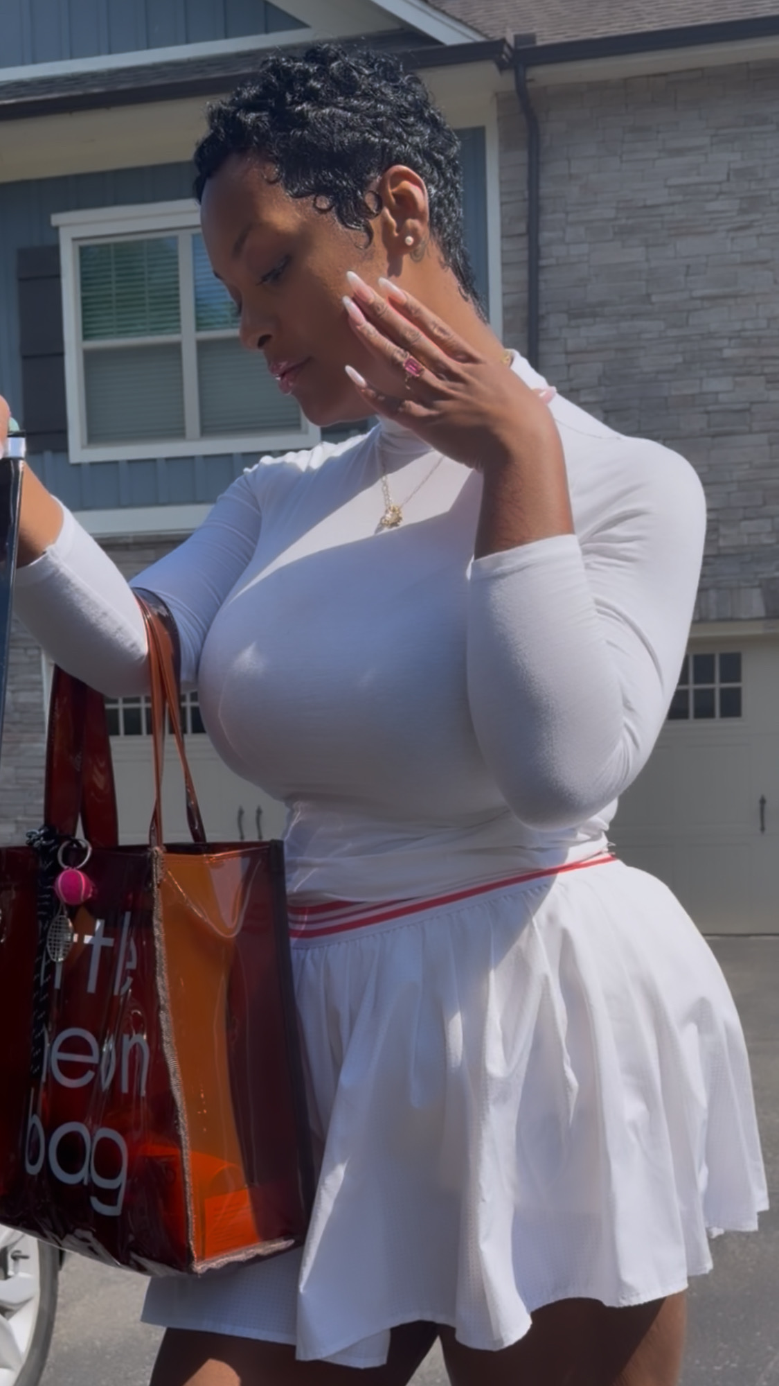 Woman in white dress holding shopping bag outdoors, touching her face, with house in background.