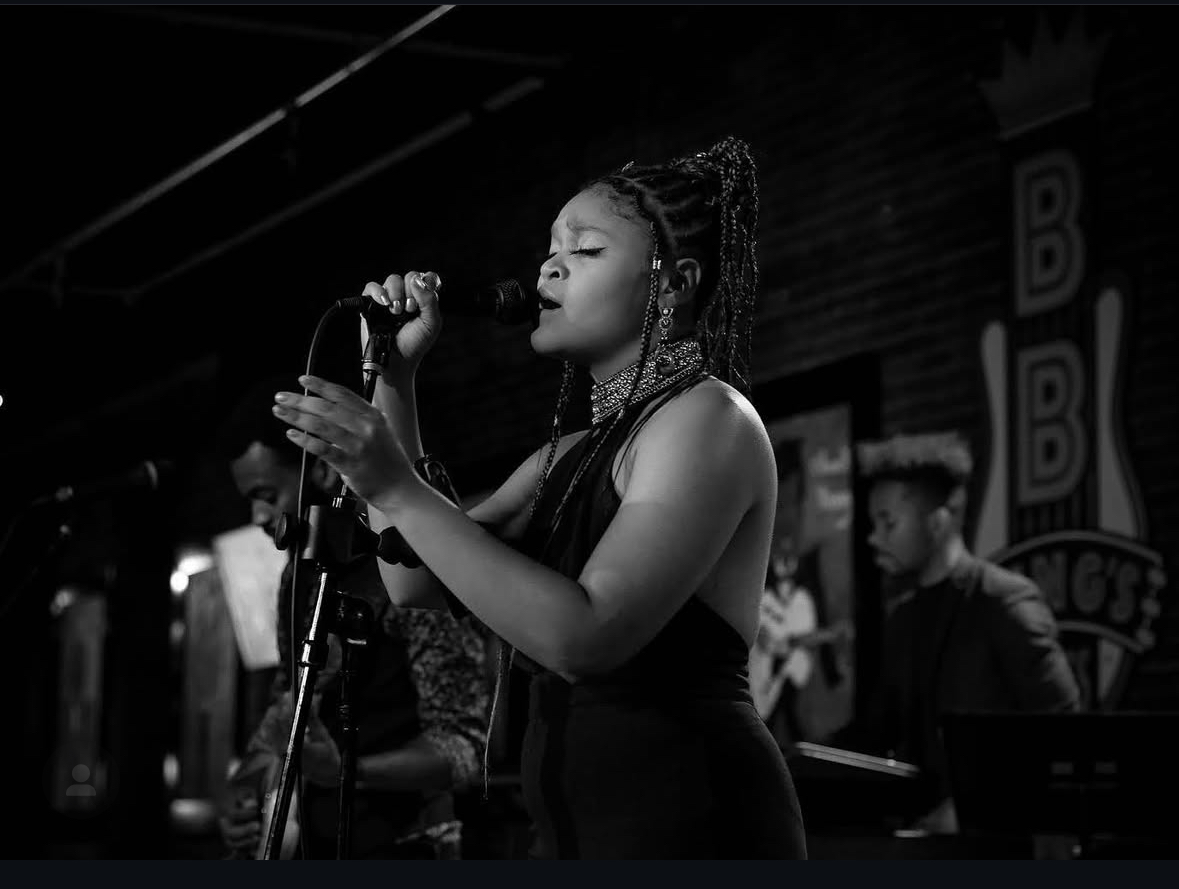 A woman singing into a microphone on stage with a band in the background, in black and white.