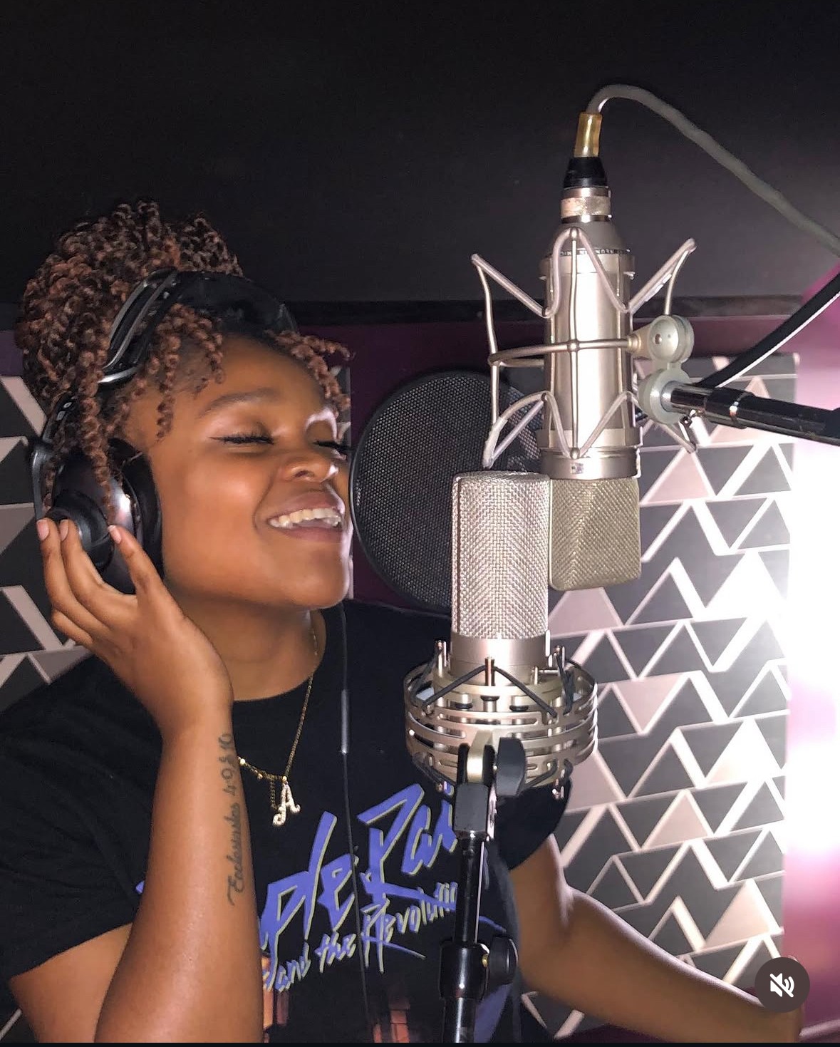 Woman in a recording studio wearing headphones, smiling, with a microphone and soundproofing panels behind her.