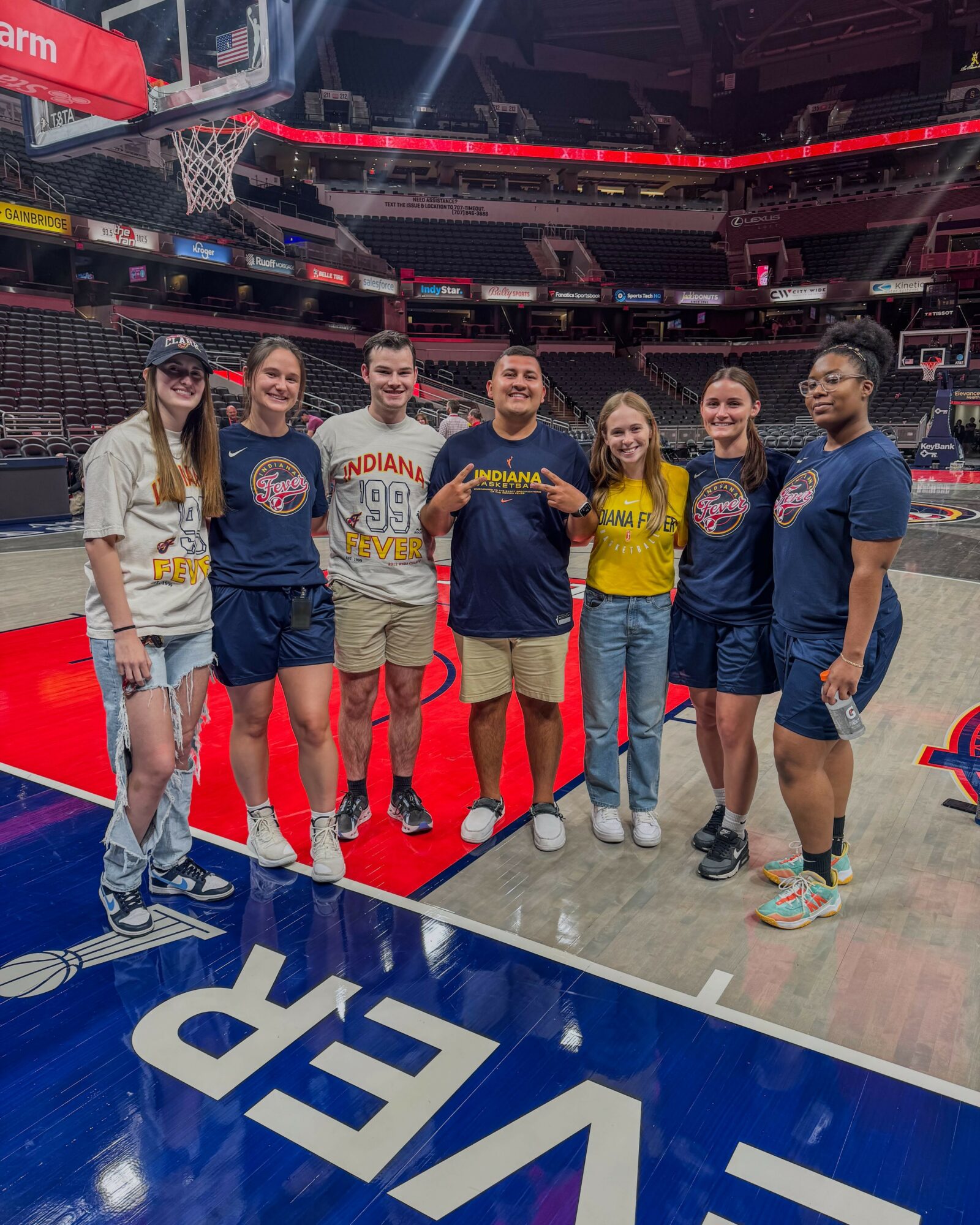 Group of seven people standing on a basketball court inside an arena, smiling, with a basketball hoop in the background.