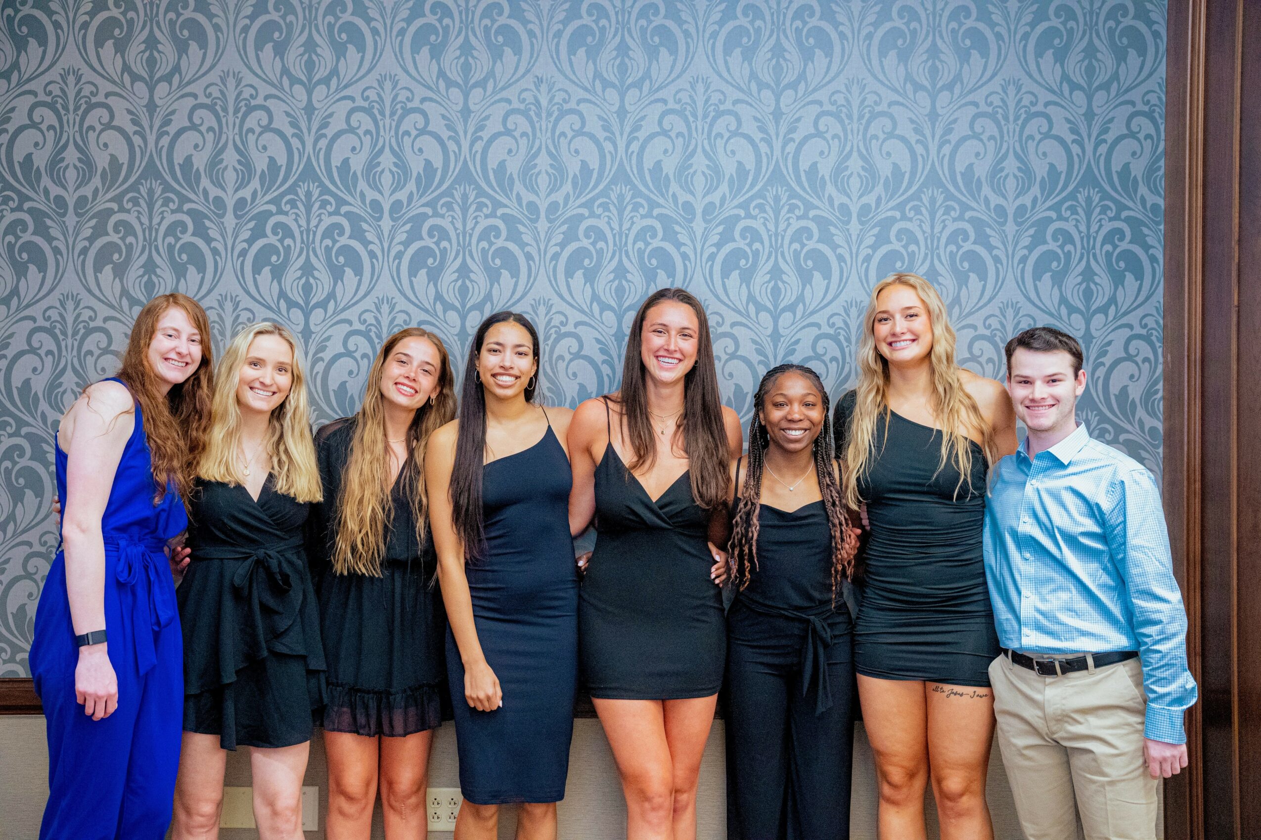 Group of seven young women and one young man standing together, smiling, in front of a patterned blue wall.