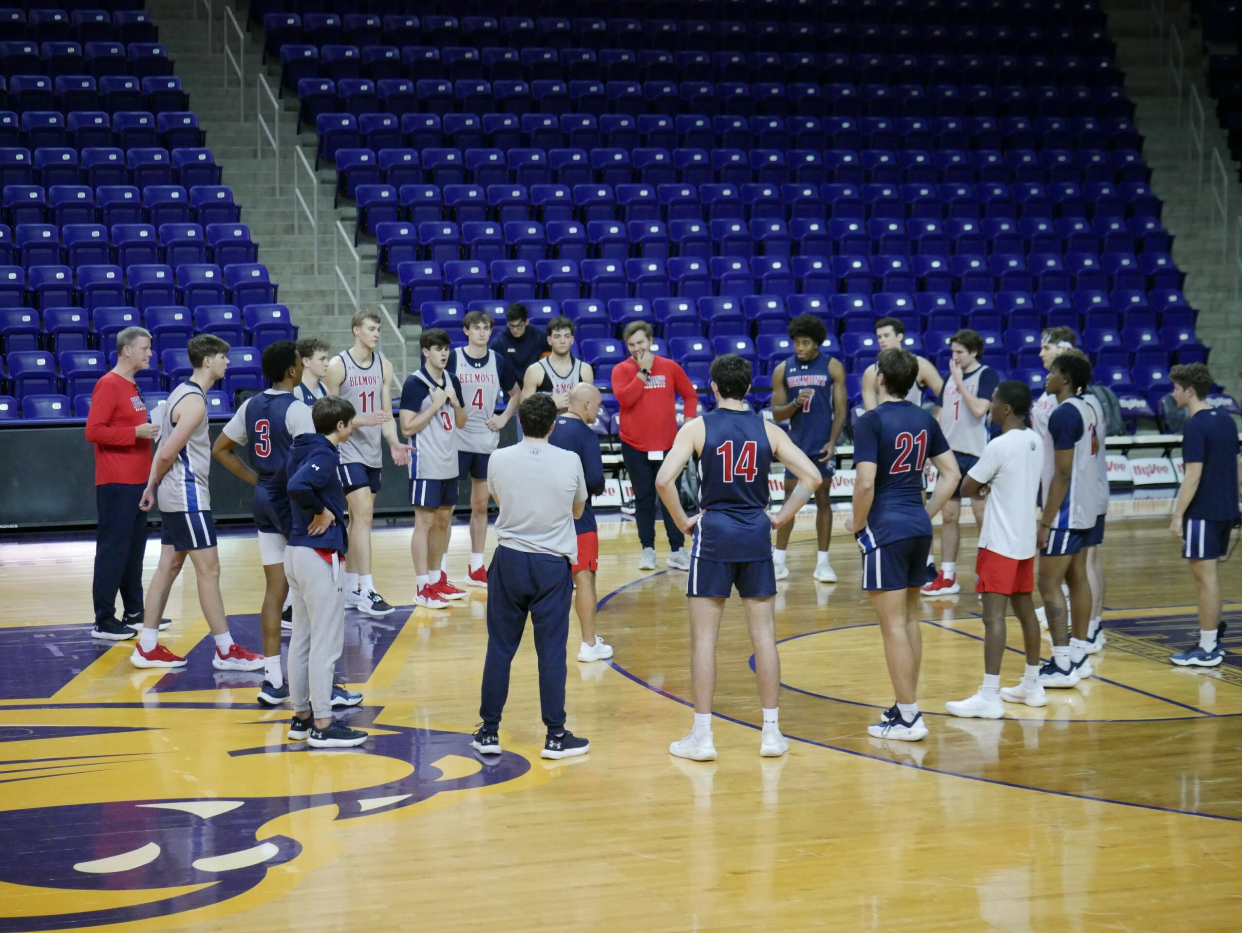 Basketball team huddles on court with coach, players in navy and white uniforms, empty blue seats in background.