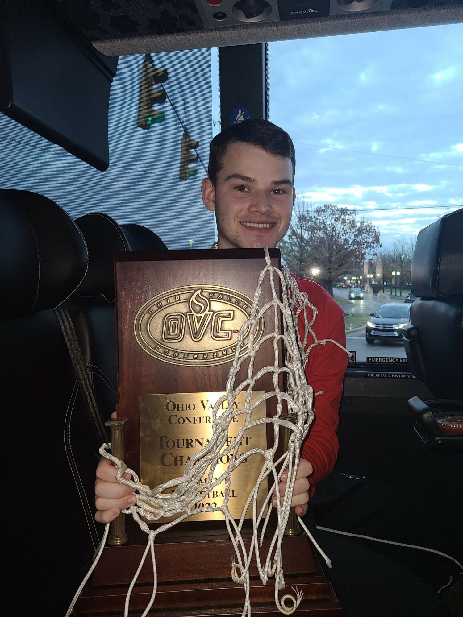 Young man holding a large award plaque inside a vehicle, with a view of a parking lot and cloudy sky outside.