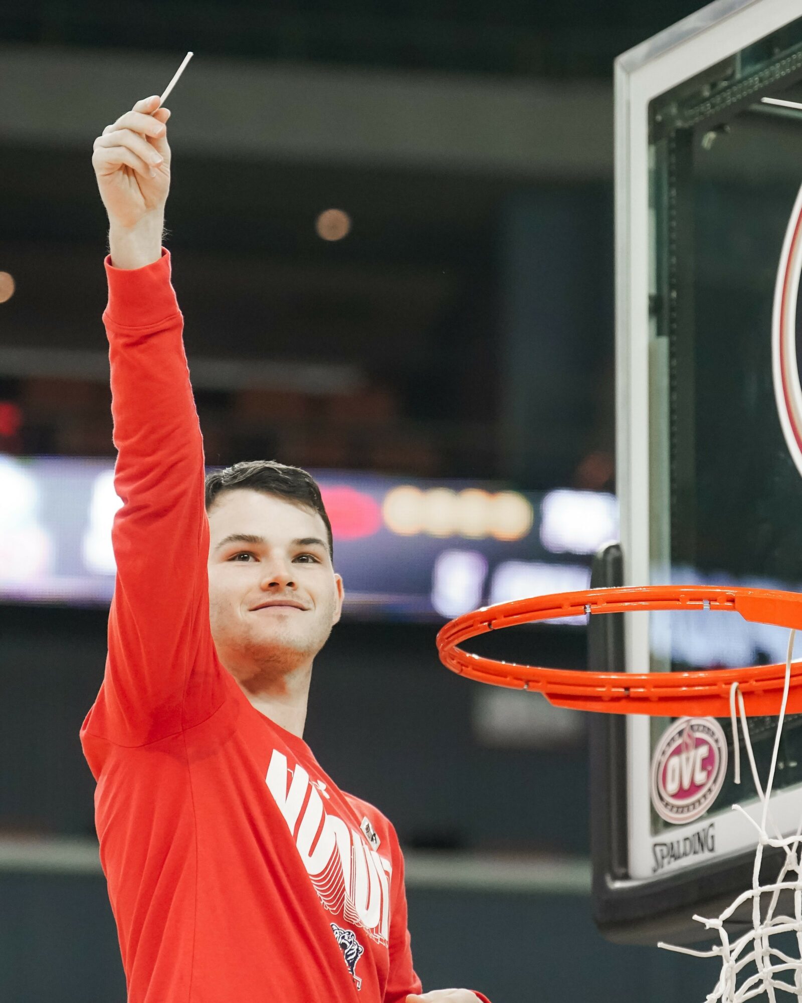 Young man in red shirt holding a stick, standing near a basketball hoop in an indoor arena.