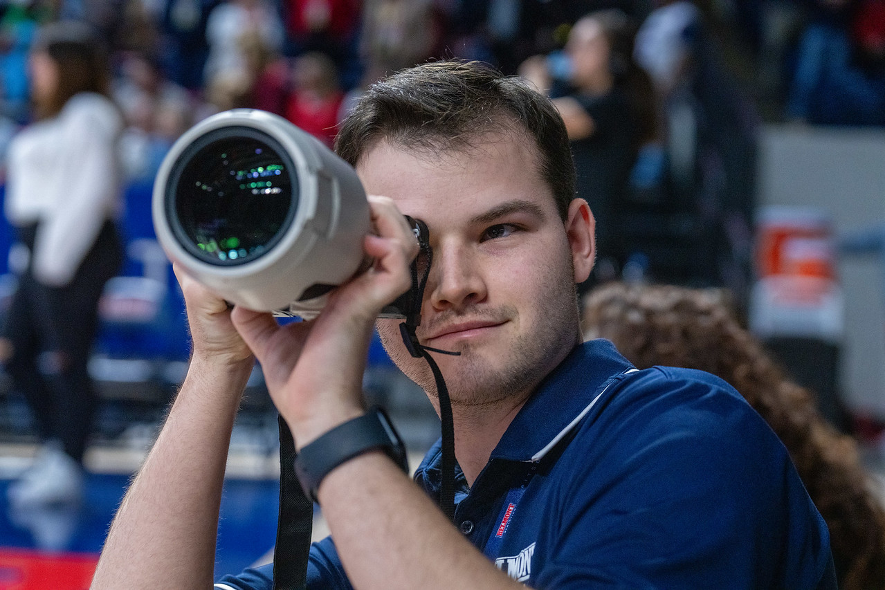 Young man looking through a telescope at an outdoor event, with a crowd in the background.