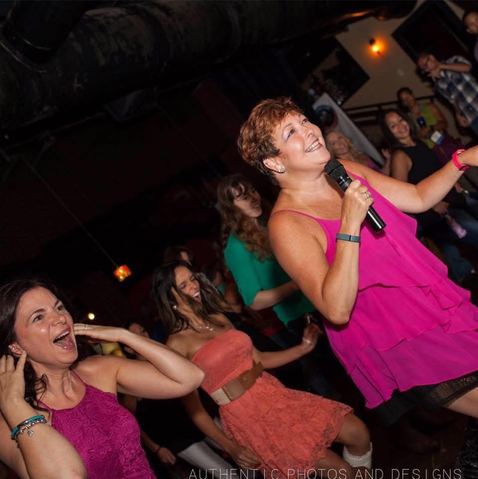 Woman in pink dress singing into microphone, smiling, with people dancing and enjoying in background.