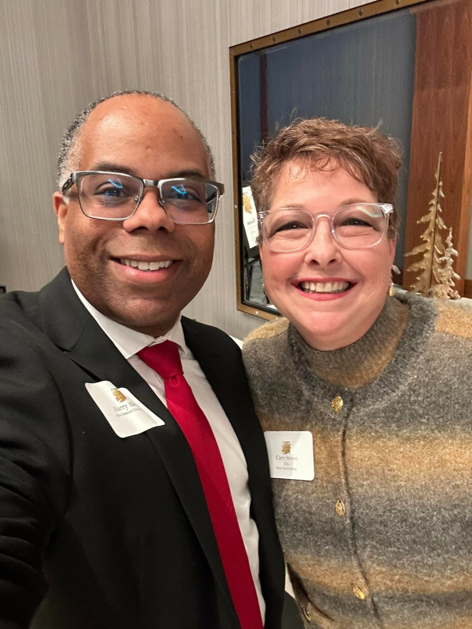 Two smiling people wearing glasses and name tags, standing close together indoors with a window behind them.