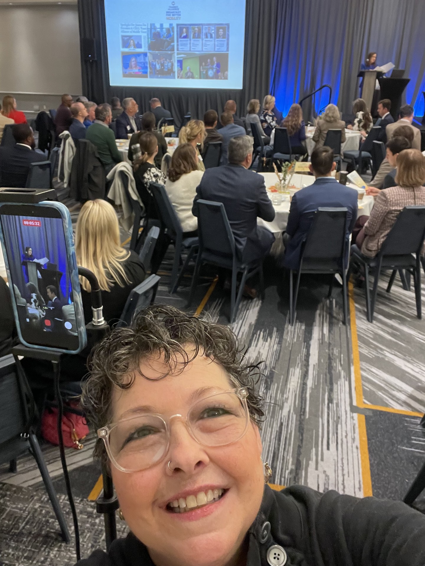 Person taking a selfie at a conference with a large audience and presentation screen in the background.