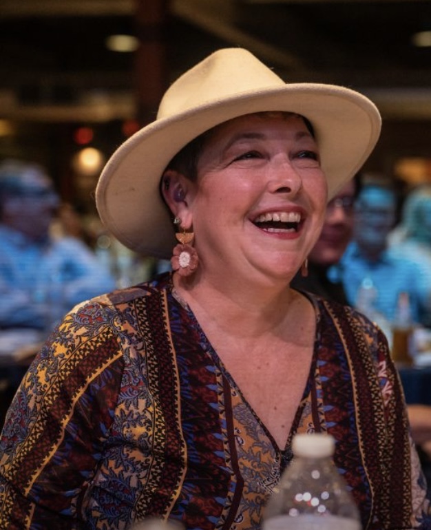 Smiling woman wearing a wide-brimmed hat and patterned clothing, with a blurred background of people indoors.