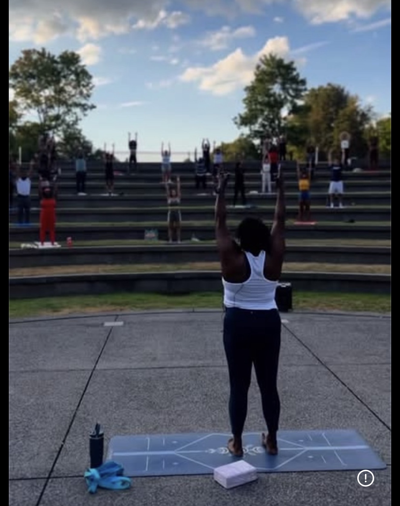 Person standing on a mat outdoors with arms raised, surrounded by others on tiered seating, trees, and sky in background.