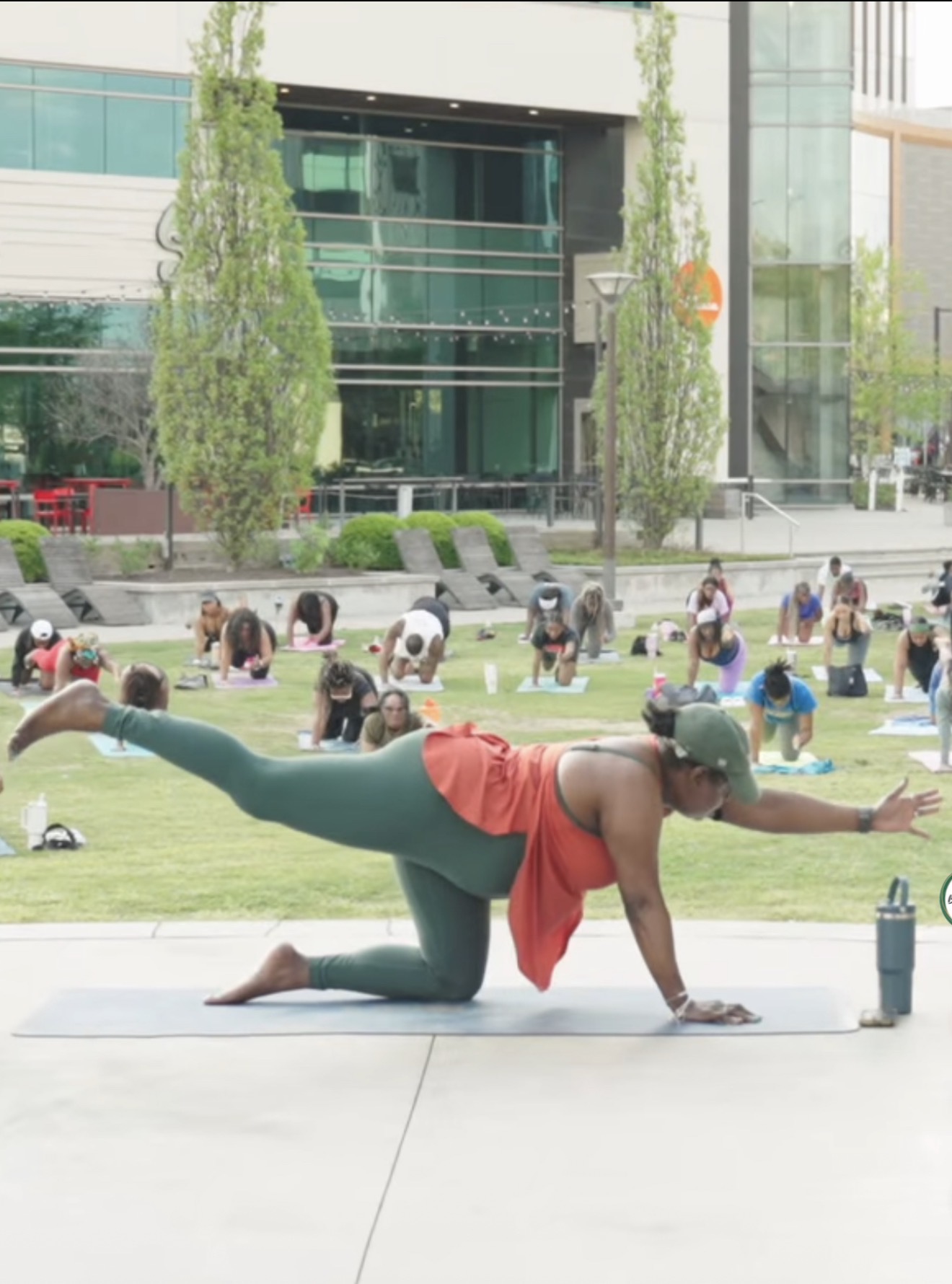 Woman practicing yoga outdoors on a mat, with a city building and trees in the background.