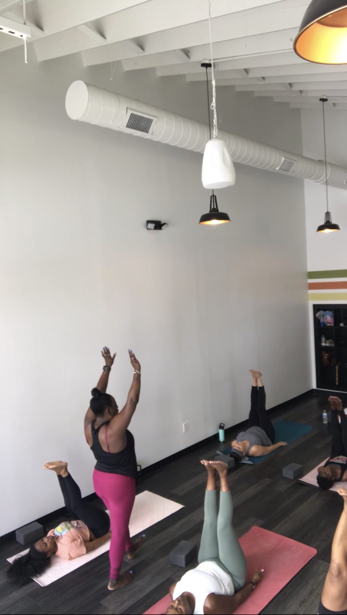 People participating in a yoga class lying on mats, with one person standing and raising arms, in a bright room.