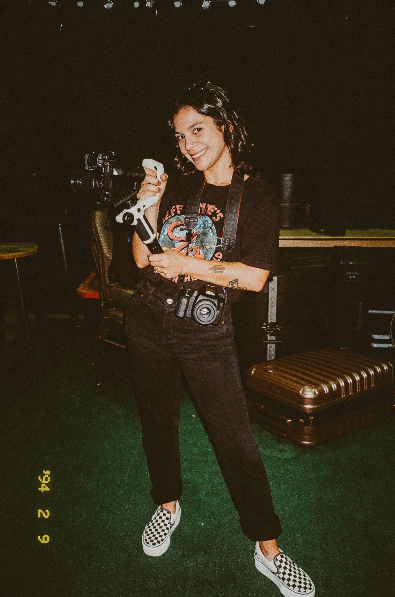 Person with curly hair holding a camera and a drink, standing indoors with dark background and a brown bench.