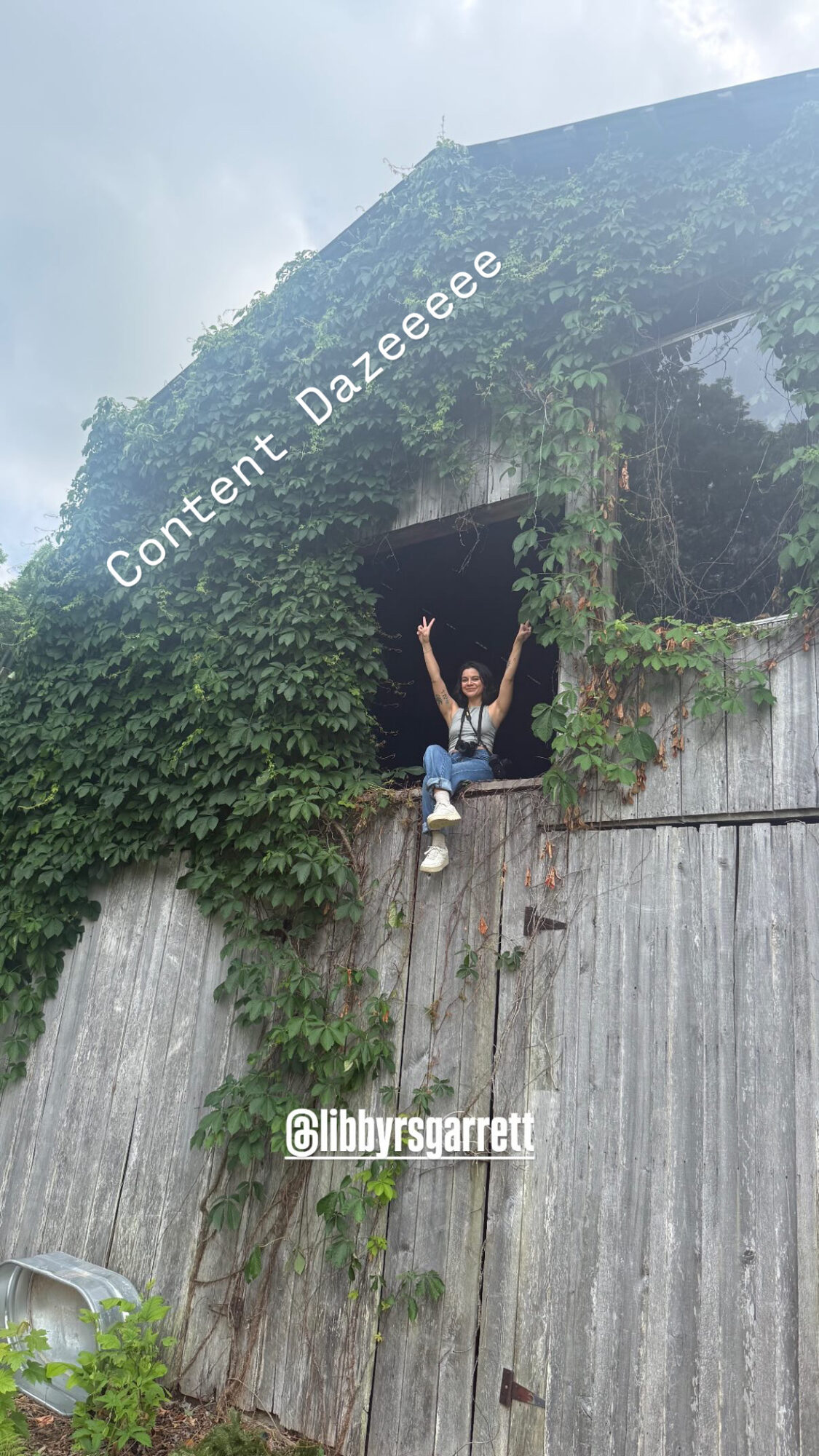 Person sitting on a ledge with arms raised, surrounded by greenery and a concrete wall, with a building covered in vines above.