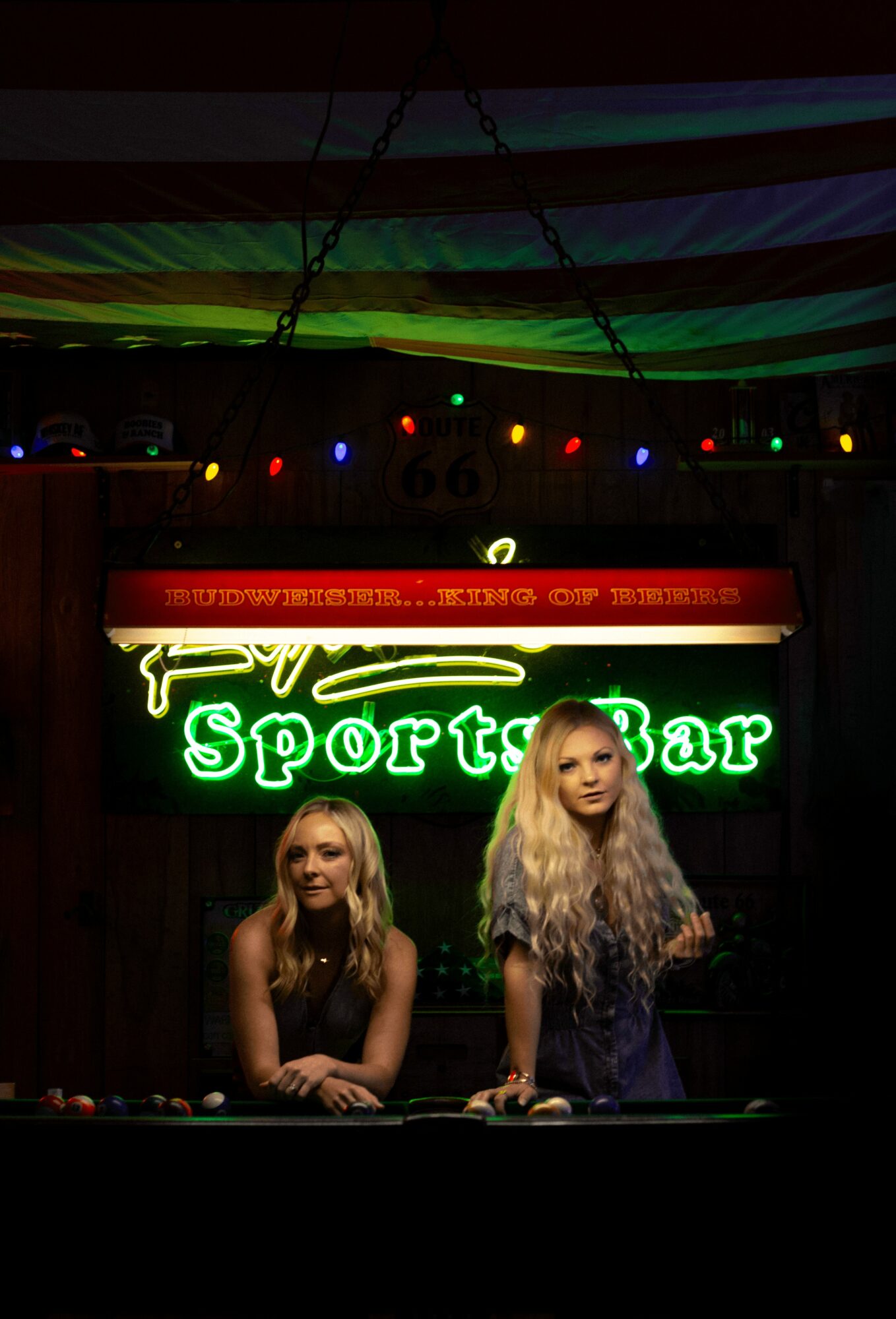 Two women with long blonde hair stand behind a bar with a neon sign reading 'Sports Bar' in green, in a dimly lit setting.